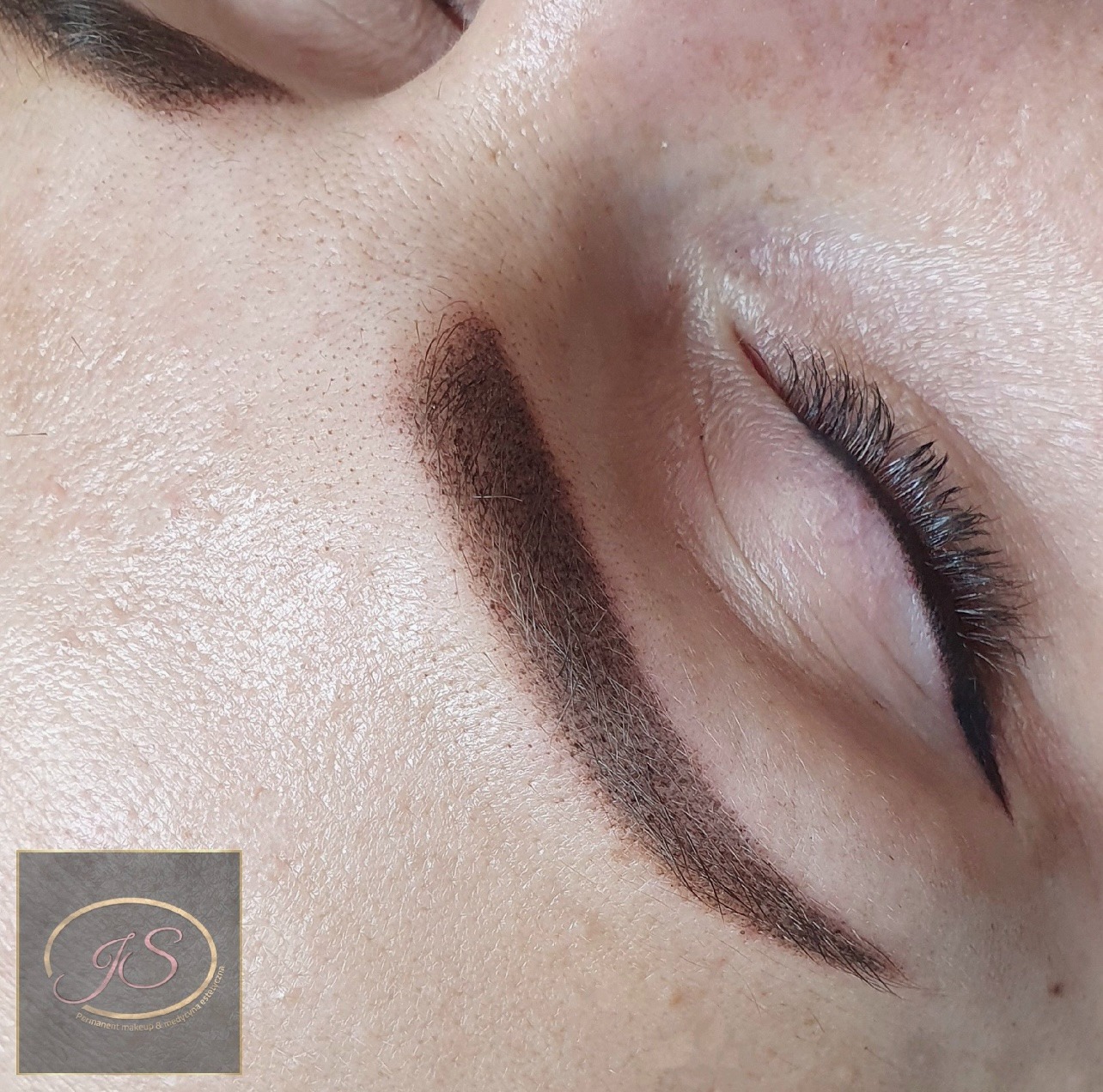 Zbliżenie na oko z widoczną brwią po zabiegu microbladingu, z delikatnie podkreślonymi rzęsami i logo firmy w rogu.
