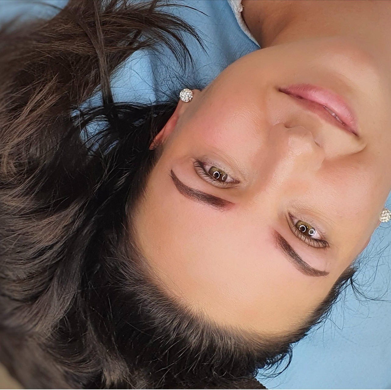 Kobieta leżąca na niebieskim podkładzie po zabiegu makijażu permanentnego brwi, widoczne kolczyki w uszach.