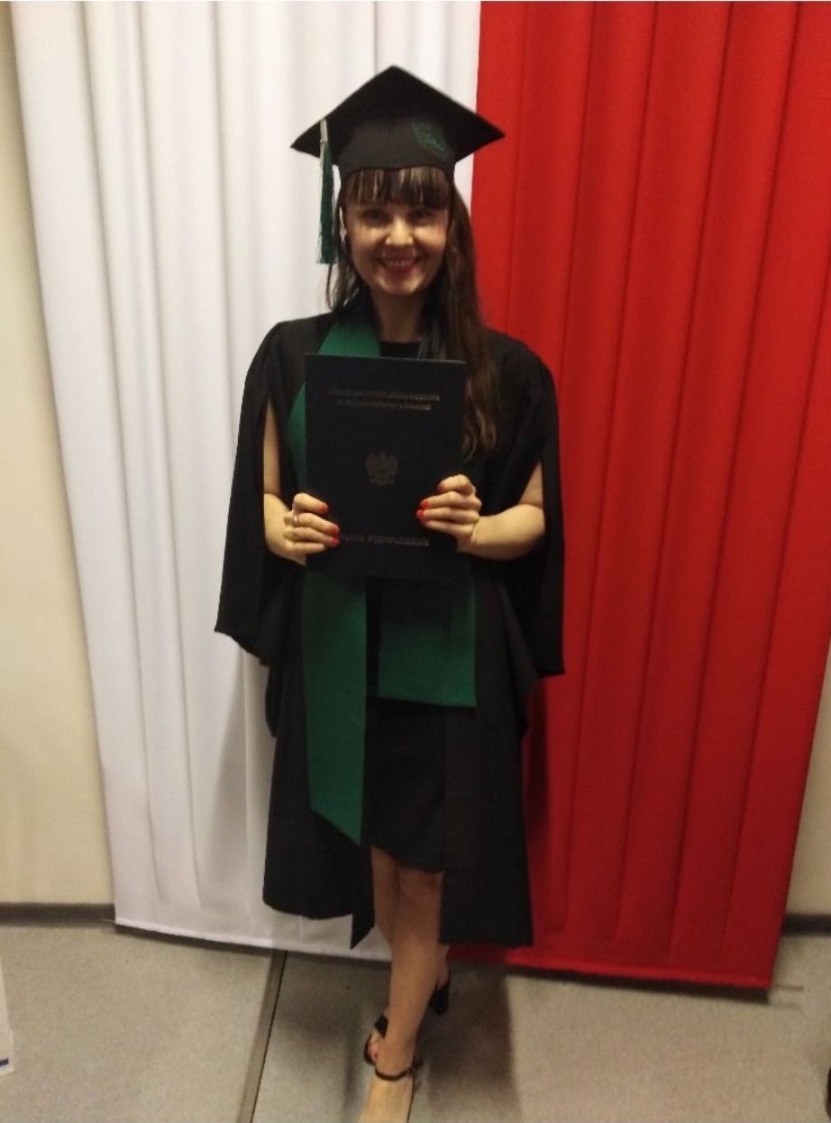 Uśmiechnięta absolwentka w todze i birecie, trzymająca dyplom na tle biało-czerwonej flagi.