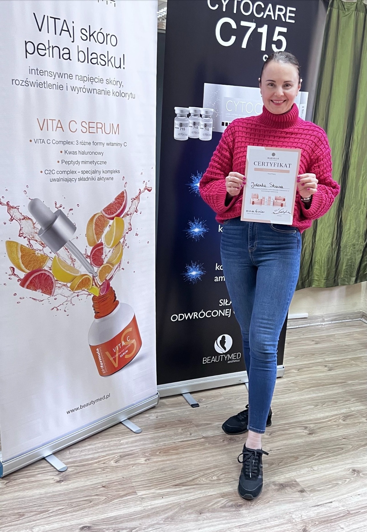 Kobieta w różowym swetrze i jeansach trzyma certyfikat na tle roll-upów reklamowych z produktami do pielęgnacji skóry.