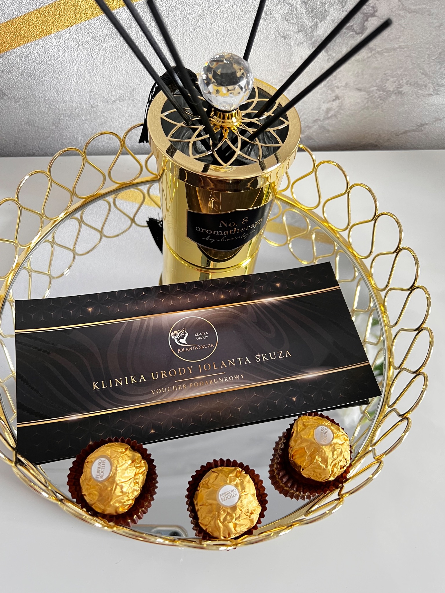 Złota taca z lustrzanym dnem, na której znajduje się czarno-złoty voucher podarunkowy Kliniki Urody Jolanta Skuza, złoty dyfuzor zapachowy z czarnymi patyczkami i kryształową gałką oraz trzy...