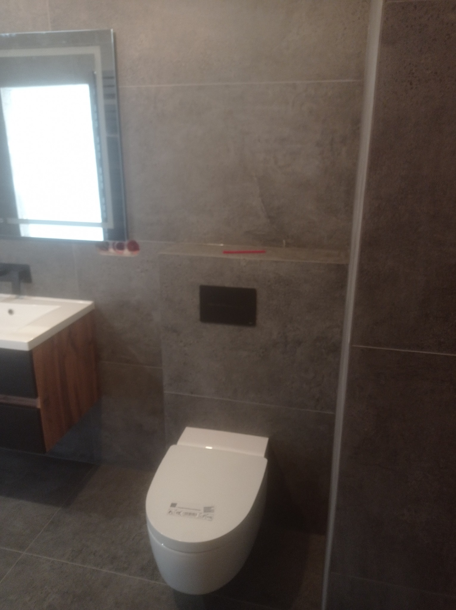 Nowoczesna łazienka z szarymi płytkami, wisząca toaleta z zabudowaną spłuczką, umywalka z drewnianą szafką i lustro z oświetleniem.