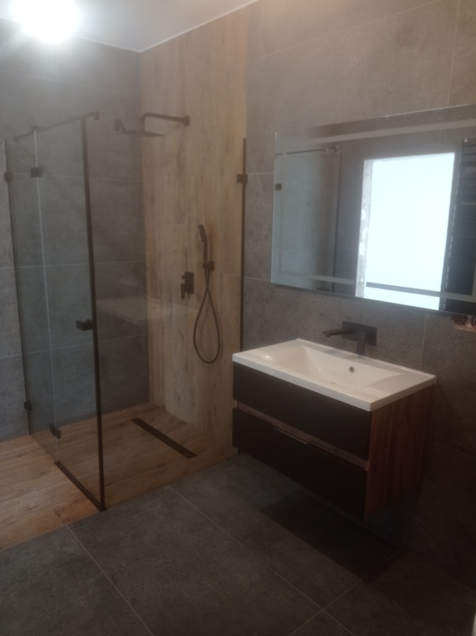 Nowoczesna łazienka z kabiną prysznicową typu walk-in z czarnymi ramami, armaturą w kolorze czarnym, umywalką z szafką w odcieniach brązu i szarości oraz dużym lustrem.