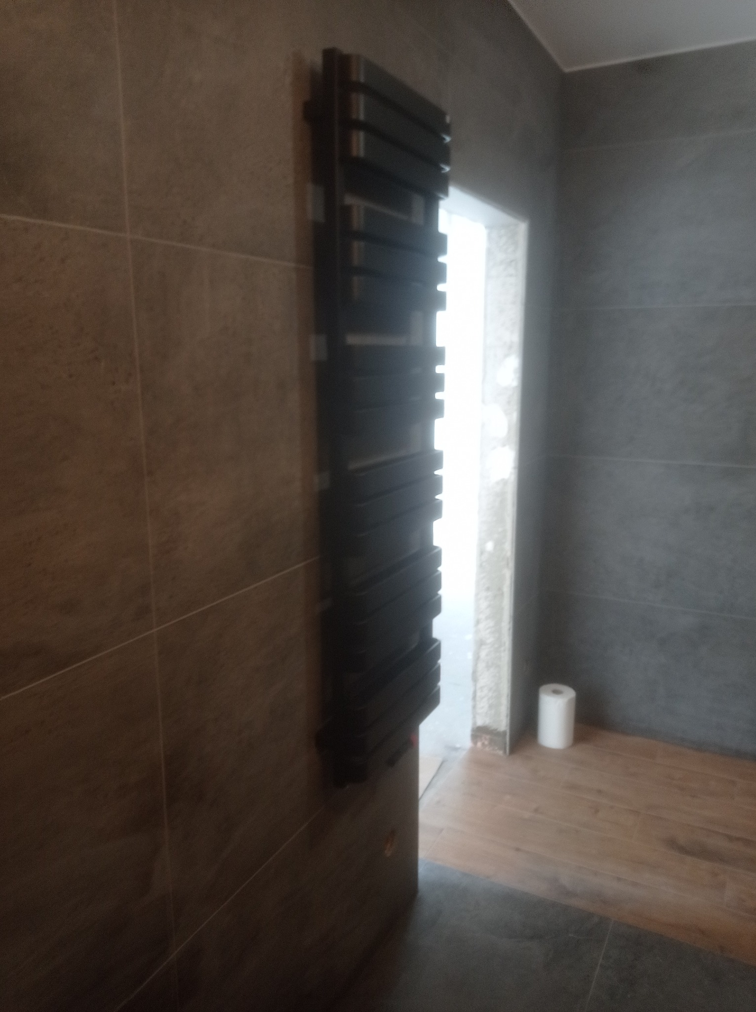 Czarny, drabinkowy grzejnik łazienkowy zamontowany na ścianie z szarych płytek w trakcie remontu łazienki, widoczne niedokończone wykończenia i rolka papieru toaletowego w tle.