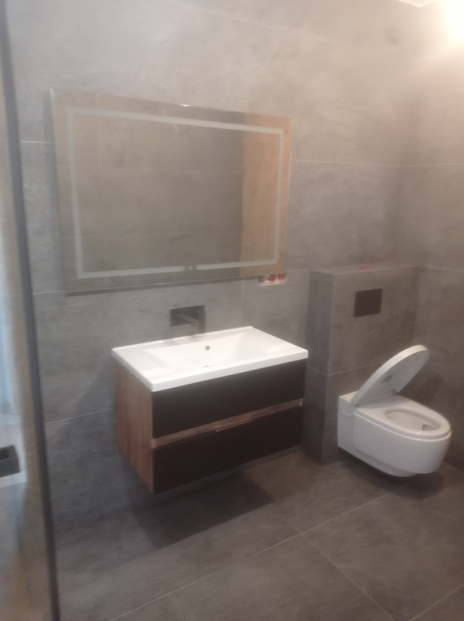 Nowoczesna łazienka wykończona szarymi płytkami, z podwieszaną szafką umywalkową z drewnopodobnym frontem i podświetlanym lustrem oraz podwieszaną toaletą z otwartą klapą.