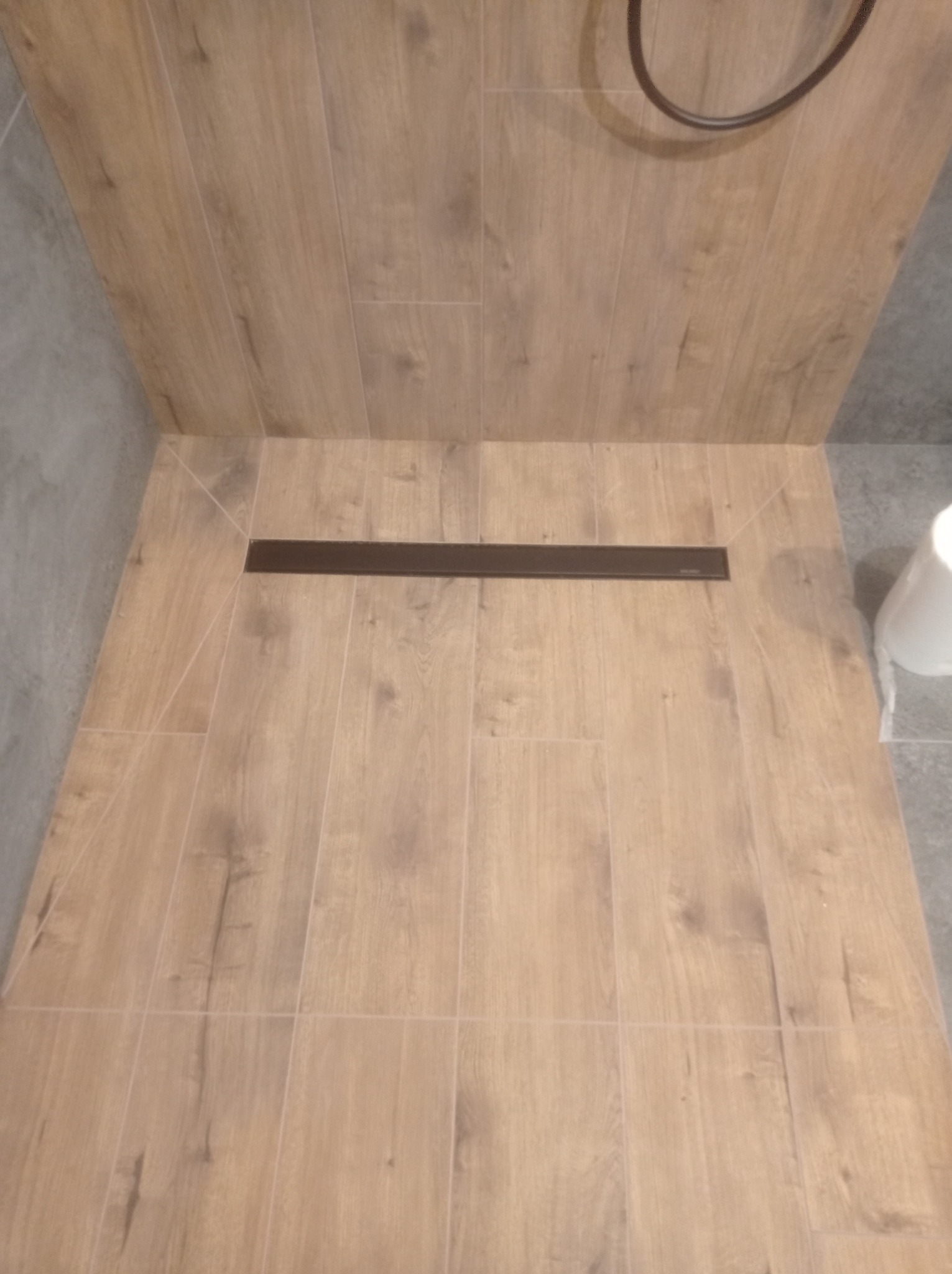 Wykończona podłoga w kabinie prysznicowej z odpływem liniowym, płytki imitujące drewno, szara ściana w tle.