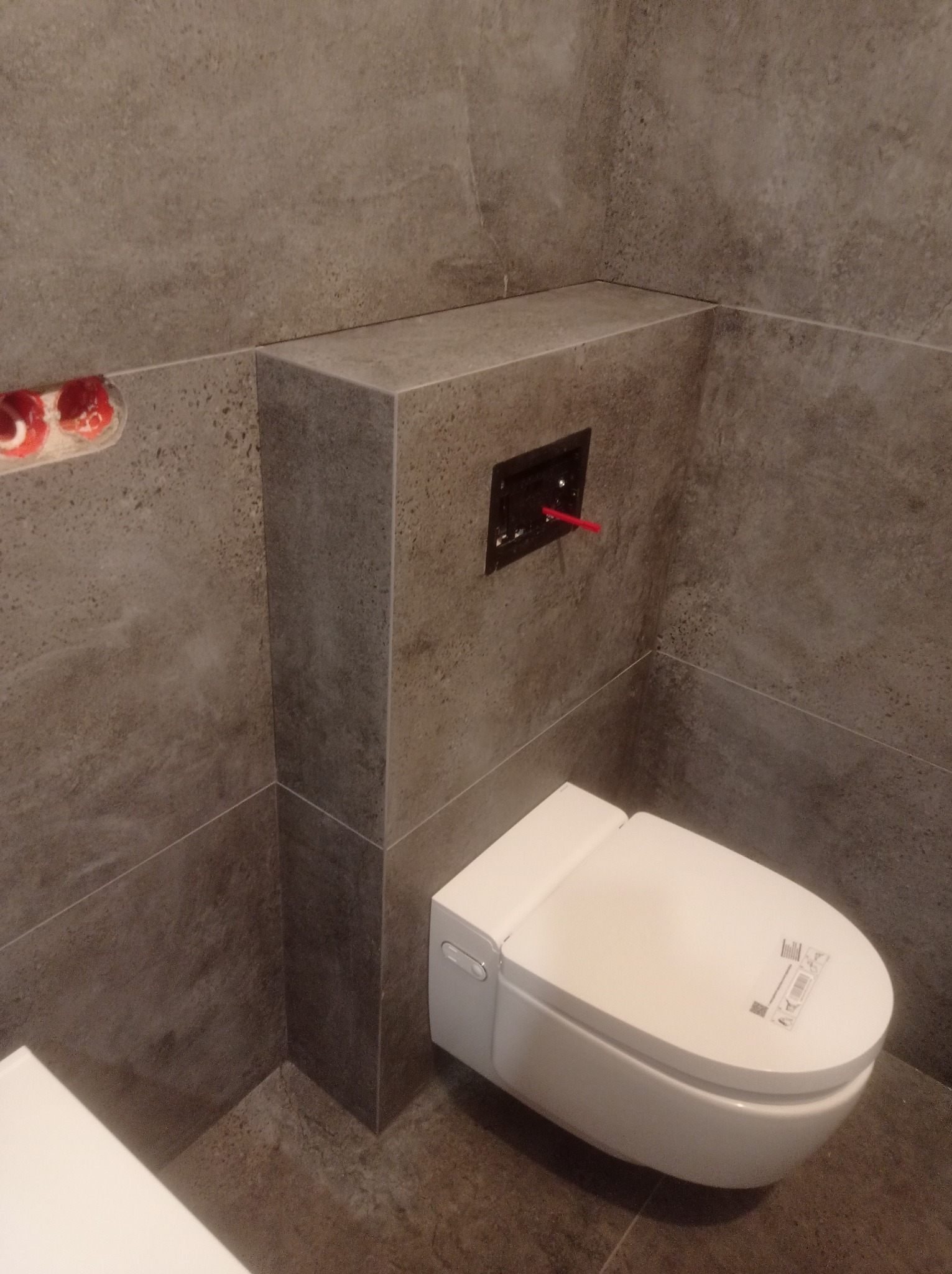 Zamontowana, biała toaleta myjąca z ukrytym stelażem podtynkowym, obudowana płytkami imitującymi kamień. Widoczne przyłącza wodne i kanalizacyjne.