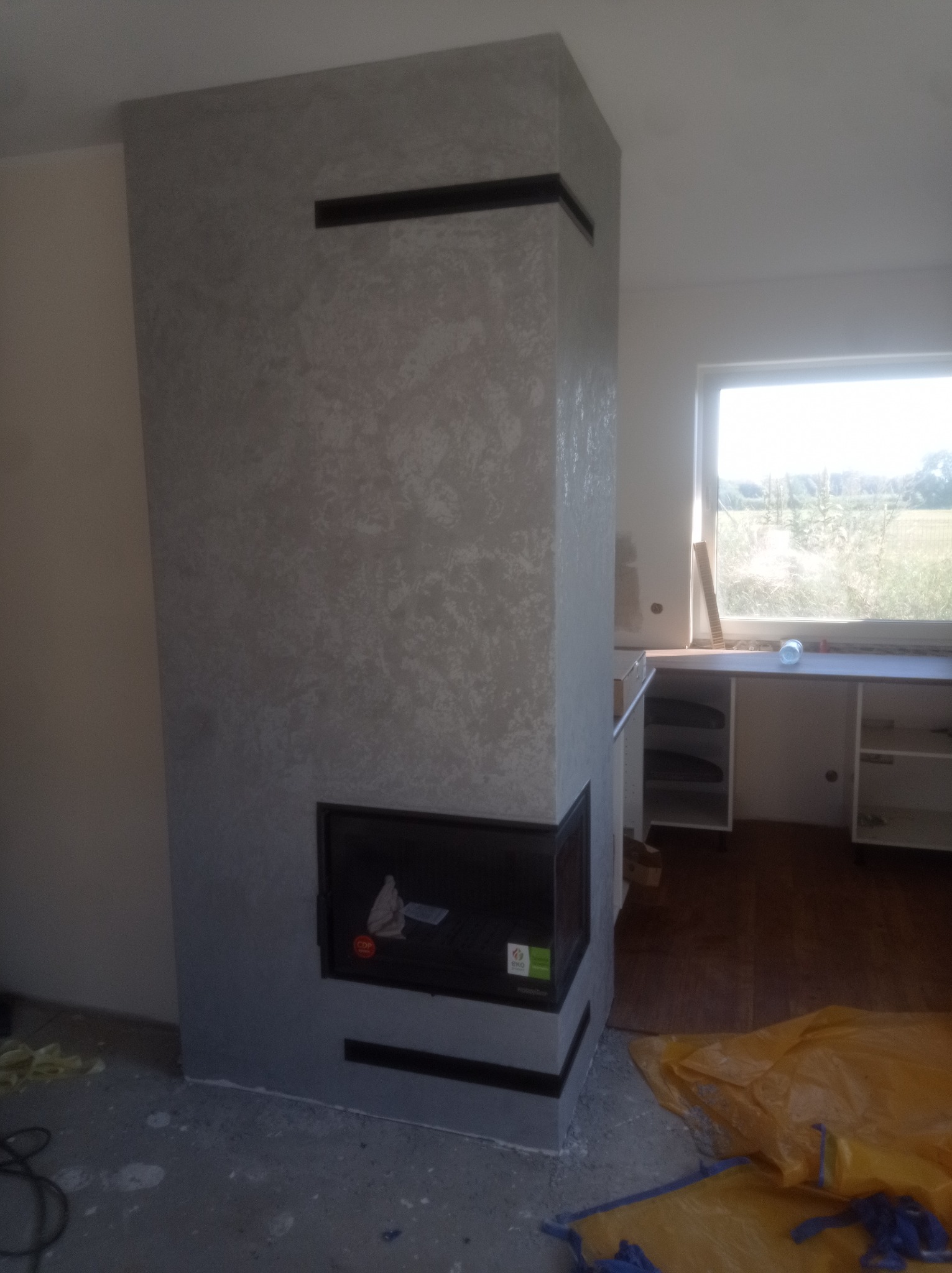 Wykończony kominek z szarą, teksturowaną obudową i czarnymi akcentami, widoczny w surowym wnętrzu w trakcie prac wykończeniowych, z widocznymi elementami zabezpieczającymi podłogę.