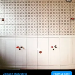 Ściana łazienki z ułożonymi jasnymi płytkami z delikatnym wzorem, poniżej biała lamperia z panelami i wystającymi rurami instalacji wodnej przed montażem armatury.