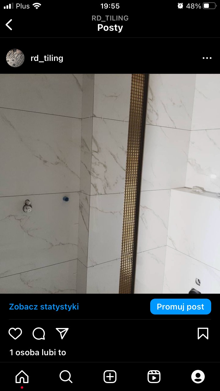 Ściana łazienki wyłożona białymi płytkami imitującymi marmur, z pionowym pasem dekoracyjnej złotej mozaiki pomiędzy płytkami, widoczne przyłącza hydrauliczne.