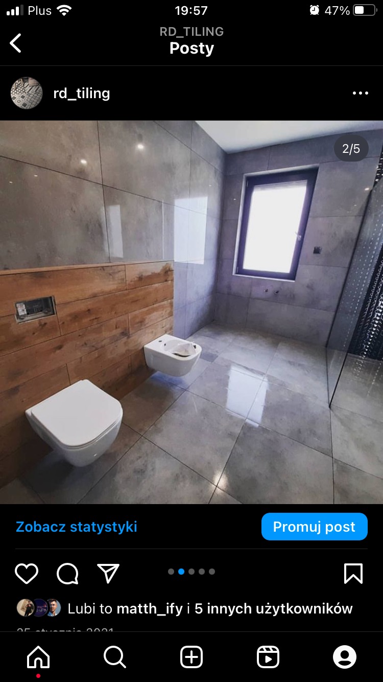 Nowoczesna łazienka z szarymi płytkami imitującymi beton, drewnianą ścianką z ukrytym stelażem WC oraz wiszącą miską i bidetem.