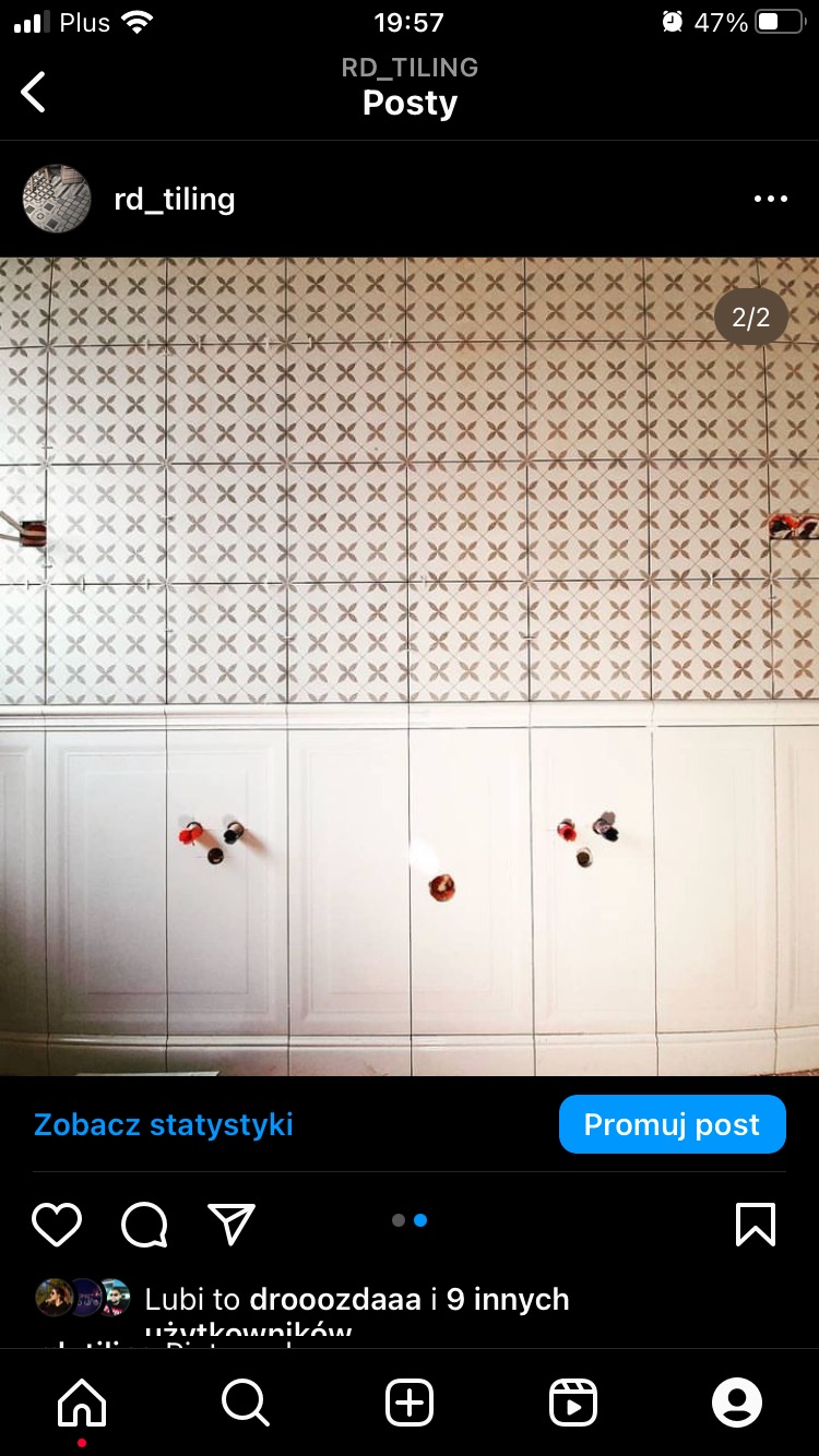 Ściana łazienki z ułożonymi jasnymi płytkami z delikatnym wzorem, poniżej biała lamperia z panelami i wystającymi rurami instalacji wodnej przed montażem armatury.