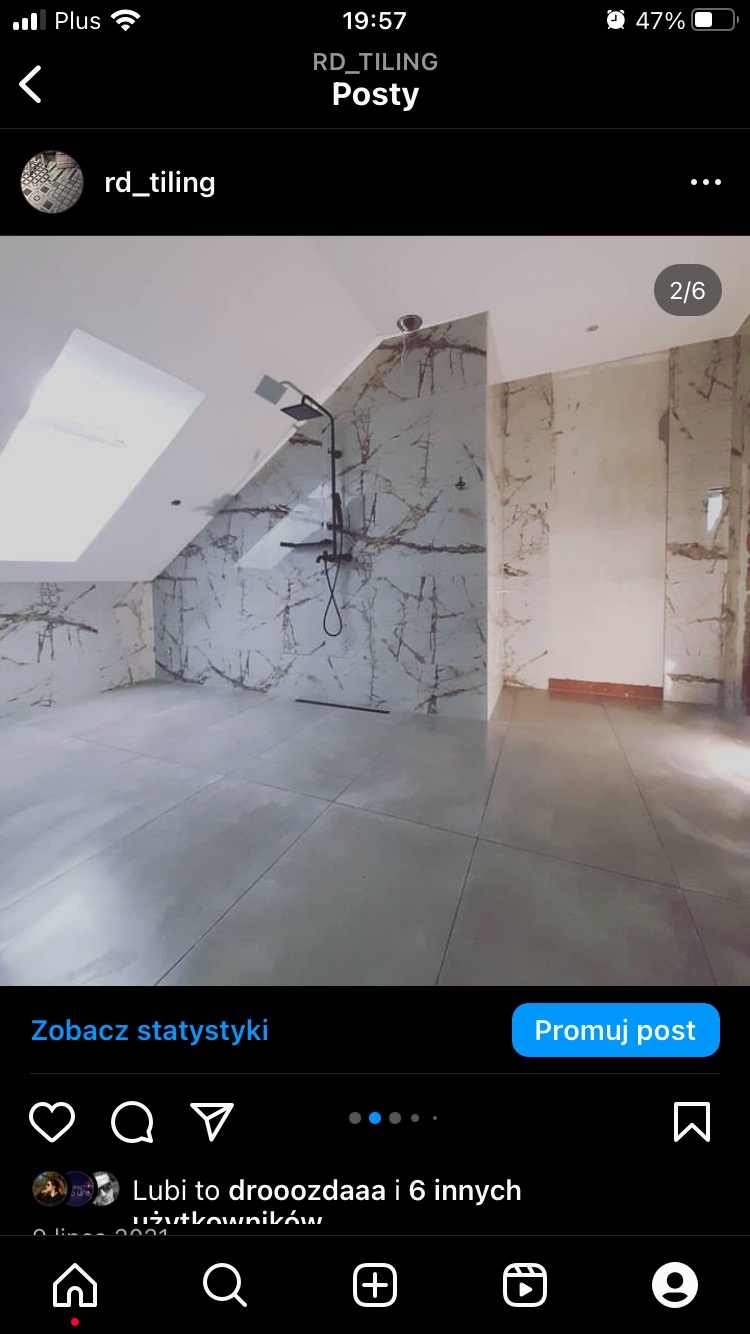 Nowoczesna łazienka na poddaszu z marmurowymi płytkami na ścianach, czarną armaturą prysznicową i szarą podłogą z dużych płyt, widok na okno dachowe.