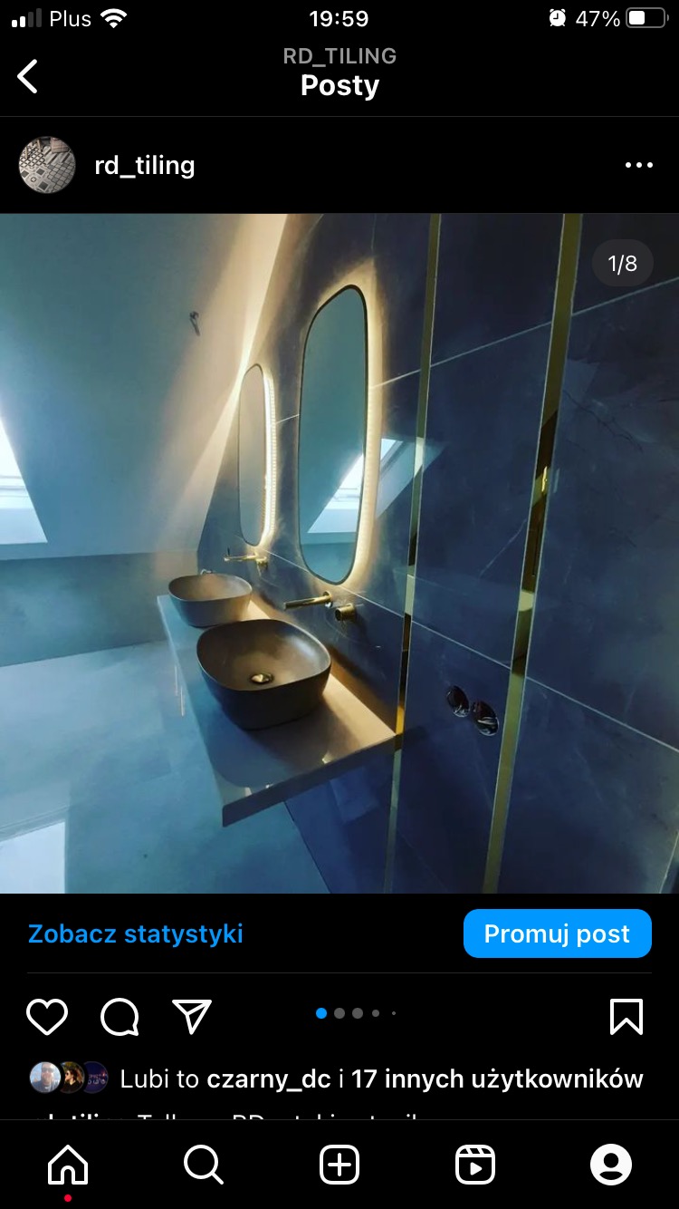 Nowoczesna łazienka na poddaszu z dwoma owalnymi umywalkami w kolorze grafitowym, podświetlanymi lustrami w złotych ramach i szarymi płytkami z złotymi listwami.