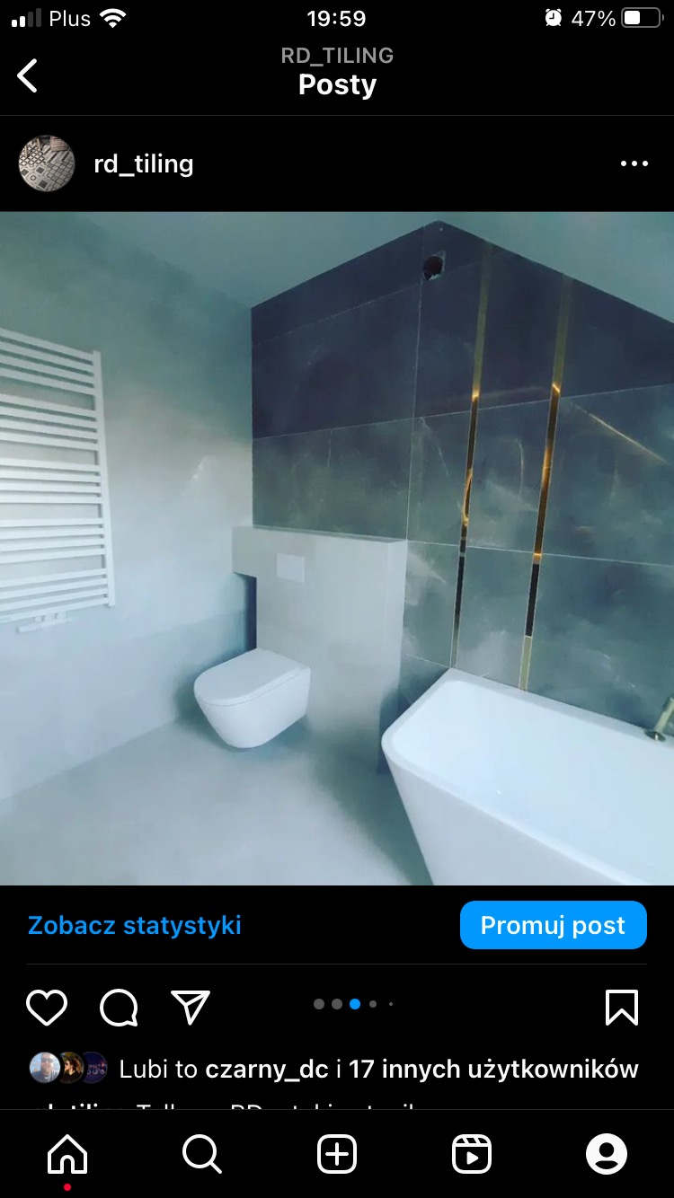 Nowoczesna łazienka z szarymi płytkami imitującymi marmur, złotymi akcentami, białą wanną wolnostojącą i podwieszaną toaletą, grzejnikiem drabinkowym.