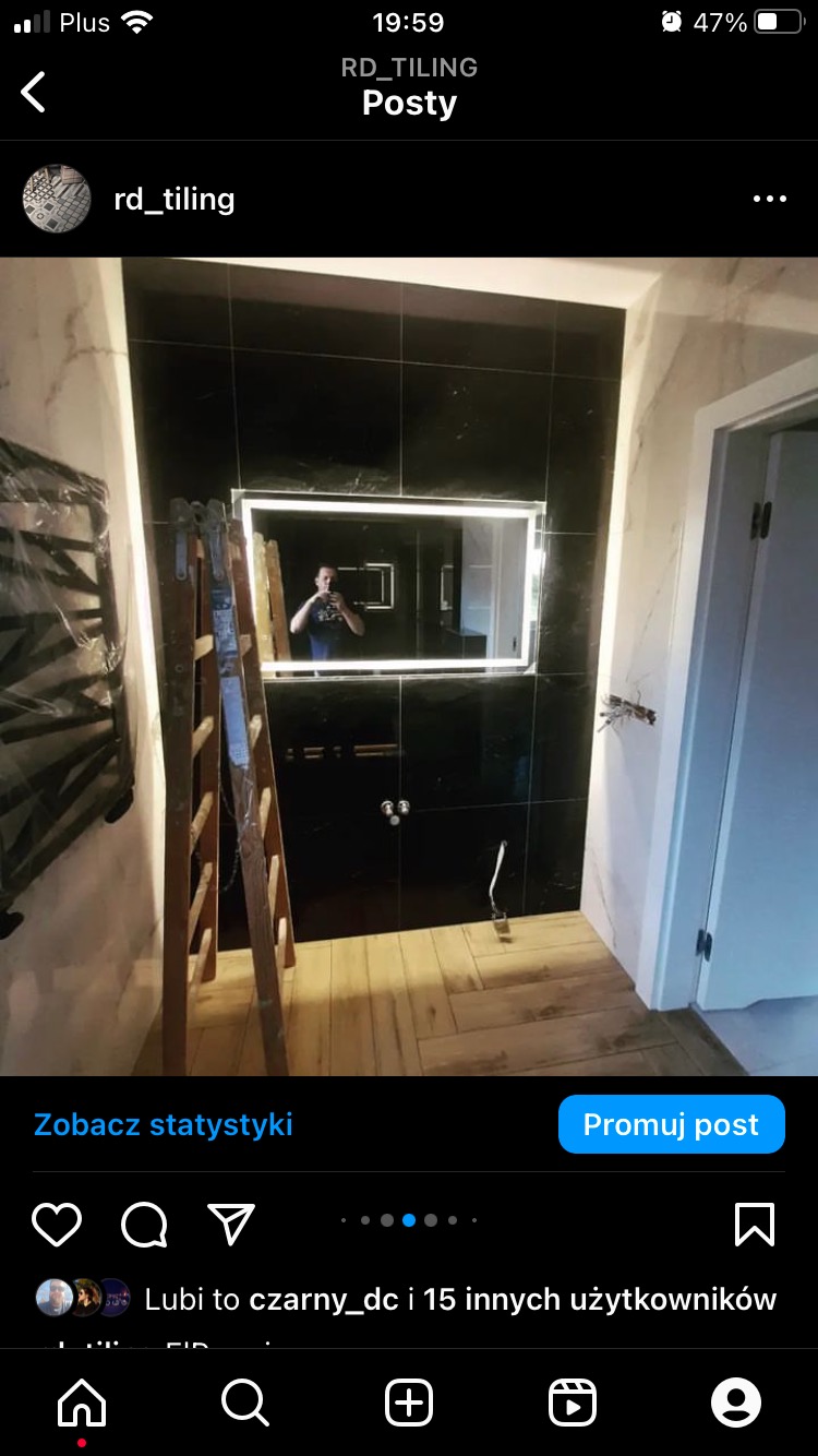 Czarne płytki w łazience z podświetlanym lustrem, drewniana drabina oparta o ścianę, widoczne narzędzia i osoba robiąca zdjęcie w odbiciu lustra.