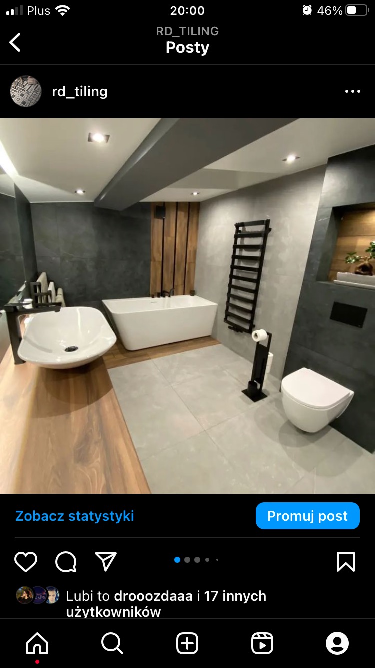 Nowoczesna łazienka z wolnostojącą wanną, umywalką nablatową na drewnianym blacie, szarą płytką na ścianach i podłodze oraz czarnymi akcentami w postaci grzejnika drabinkowego i akcesoriów...