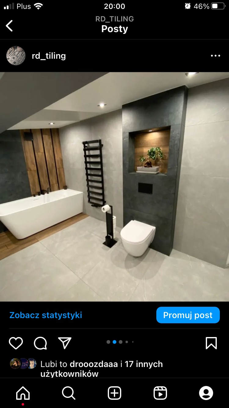 Nowoczesna łazienka z szarymi płytkami, wolnostojącą białą wanną, grzejnikiem drabinkowym i toaletą podwieszaną z zabudową z ciemnego kamienia i oświetloną wnęką z rośliną.