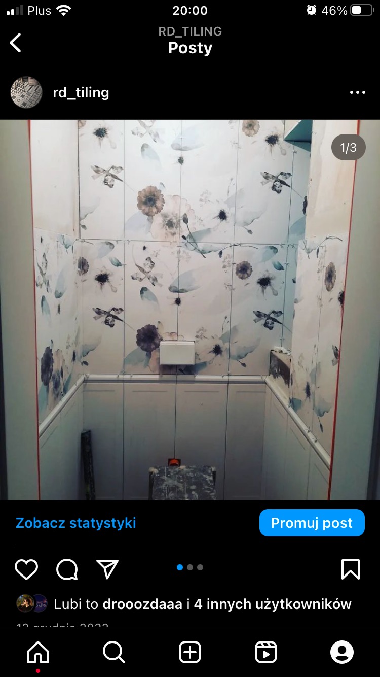 Wnętrze łazienki w trakcie wykańczania, ściany pokryte białymi płytkami z motywem kwiatowym i ważek, dolna część ścian wykończona białymi panelami, widoczny fragment toalety i narzędzia na podłodze.