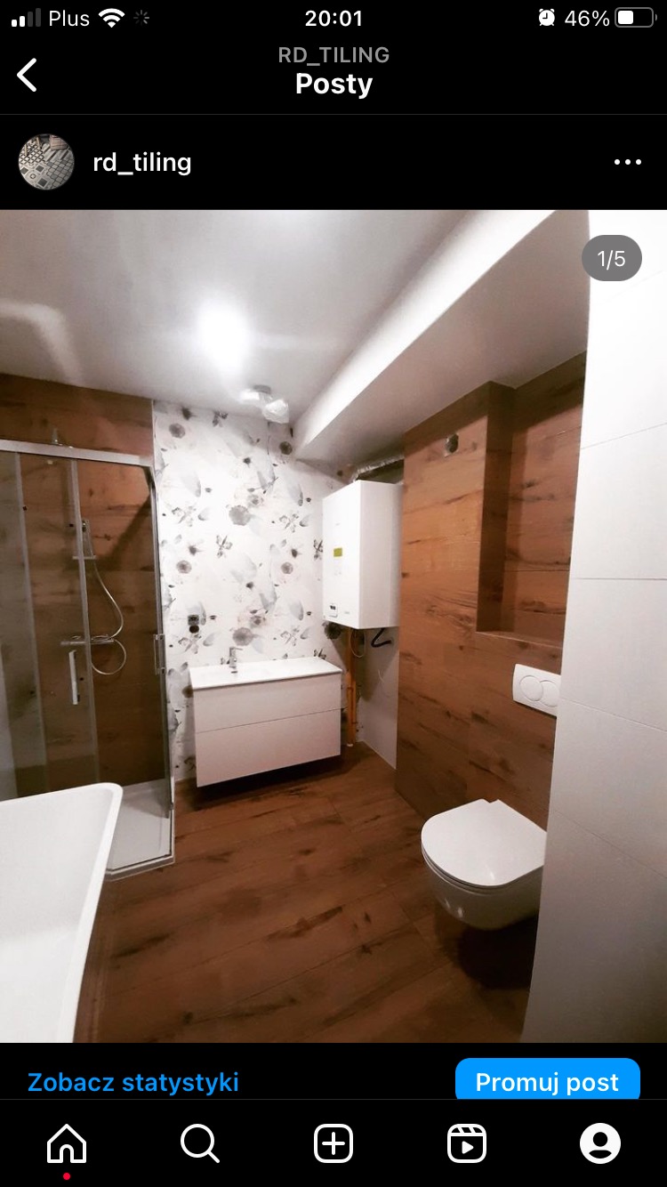 Wykończona łazienka z drewnopodobną okładziną ścienną, umywalką z szafką, kabiną prysznicową i wiszącą toaletą. Na ścianie widoczny piec gazowy.