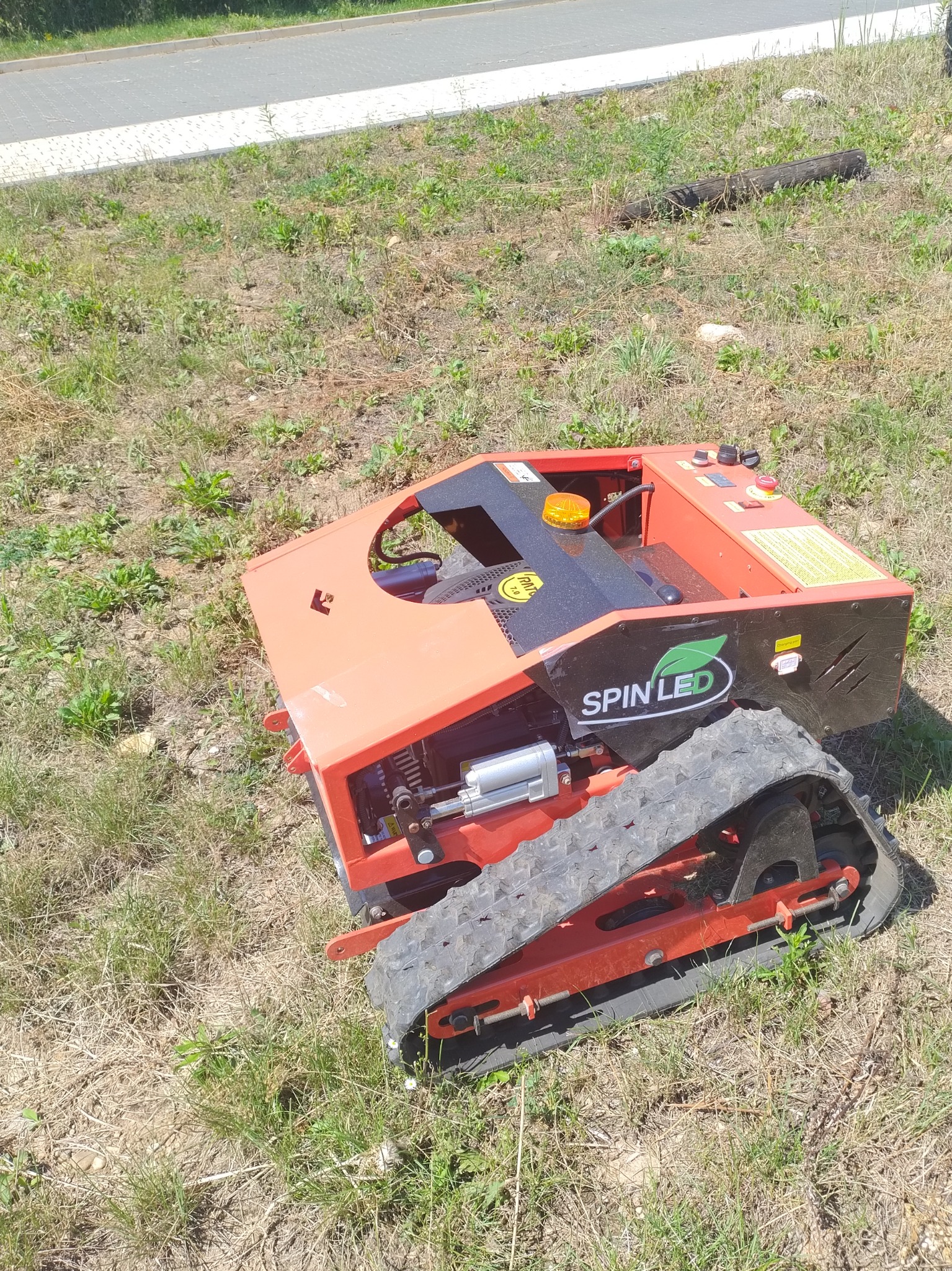 Czerwony robot koszący z napędem gąsienicowym marki Spin Led na trawiastym terenie, widoczne detale konstrukcyjne i logo.