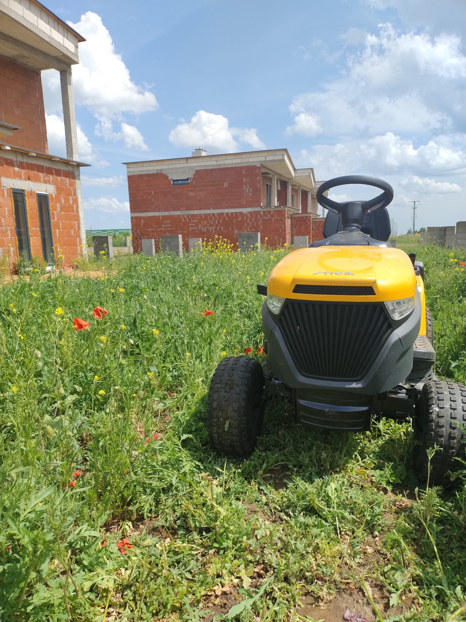 Żółta kosiarka traktorowa Stiga stoi na wysokiej, zarośniętej łące z czerwonymi makami i żółtymi kwiatami, w tle niedokończone budynki z czerwonej cegły i błękitne niebo z chmurami.