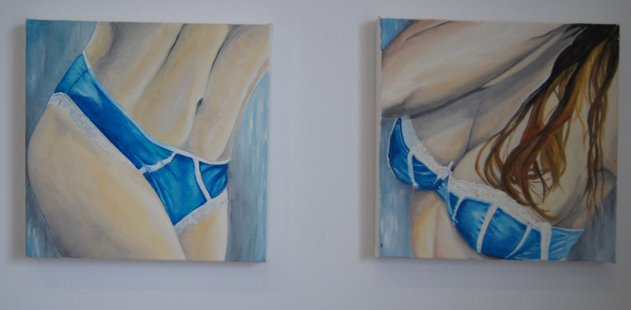 Dwa obrazy olejne przedstawiające fragmenty kobiecego ciała w błękitnej bieliźnie z koronką, zawieszone na białej ścianie.