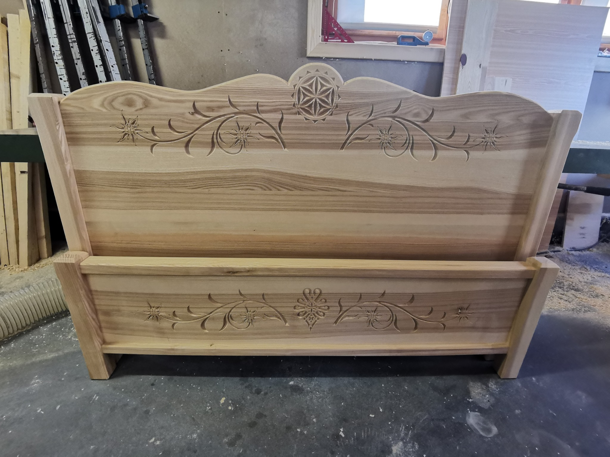 Surowe drewniane łóżko z rzeźbionymi wzorami kwiatowymi i geometrycznymi, widoczne elementy warsztatu w tle.
