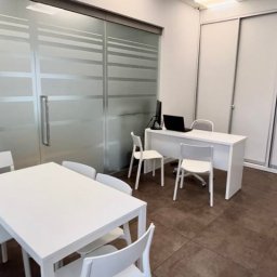 Biuro Nieruchomości BZM - Nowoczesne biuro nieruchomości z białym stołem konferencyjnym, biurkiem z laptopem i przeszkloną ścianą z poziomymi pasami. Minimalistyczny wystrój.