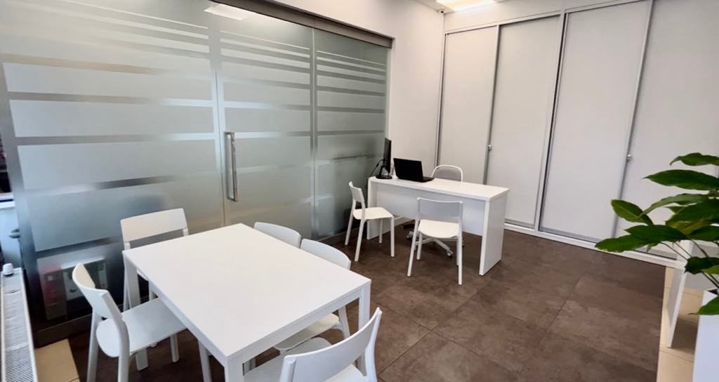 Nowoczesne biuro nieruchomości z białym stołem konferencyjnym, biurkiem z laptopem i przeszkloną ścianą z poziomymi pasami. Minimalistyczny wystrój.