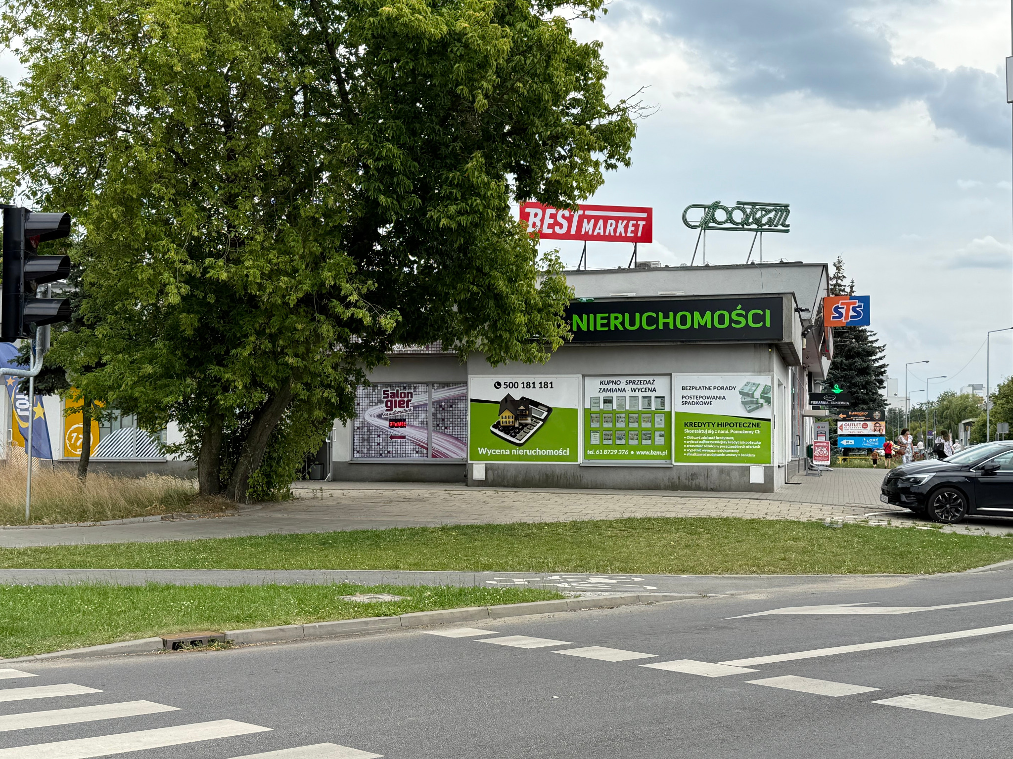 Budynek agencji nieruchomości z reklamami na elewacji, widoczny z ulicy. Na dachu szyldy sklepów. Zieleń drzew i trawników w otoczeniu. Przejście dla pieszych na pierwszym planie.