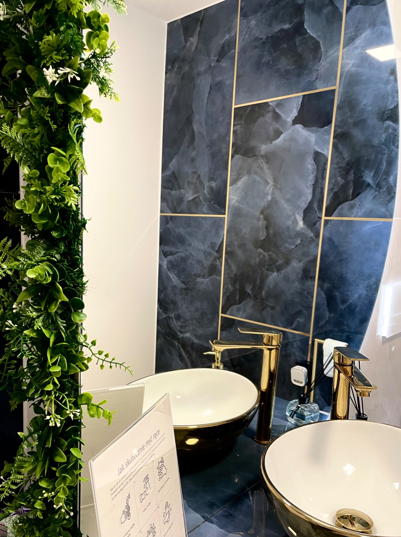 Nowoczesna łazienka z dwoma umywalkami nablatowymi w kolorze czarnym ze złotymi akcentami, złotymi bateriami, ścianą z płytek imitujących marmur z geometrycznymi złotymi wstawkami, dekoracją...