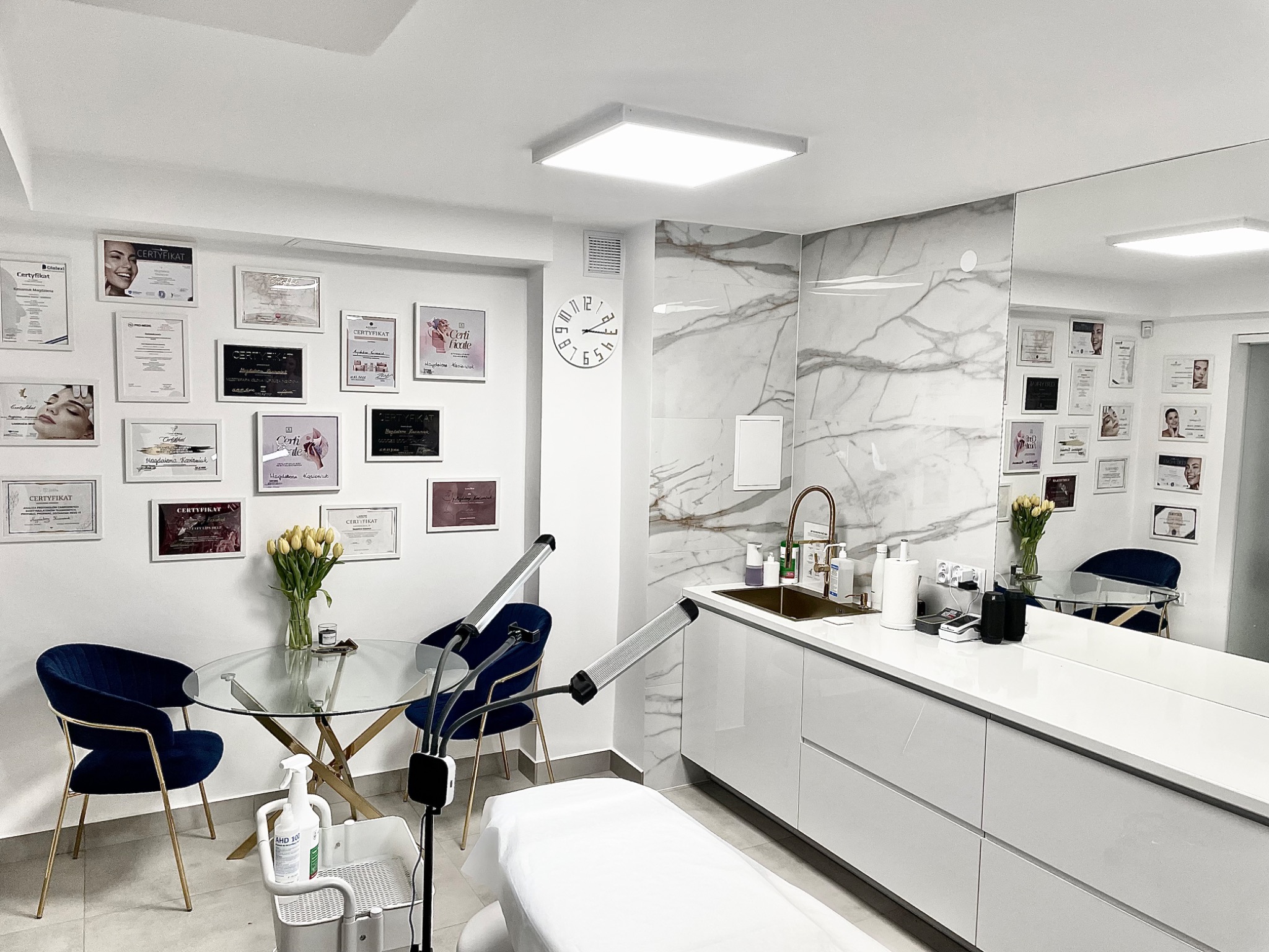 Wnętrze gabinetu z łóżkiem kosmetycznym, stolikiem z kwiatami, umywalką i ścianą z certyfikatami, oświetlone lampami punktowymi.