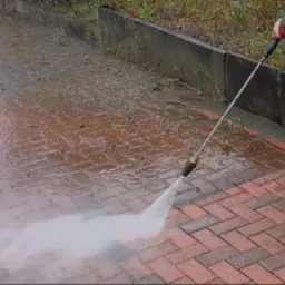 Ceglana kostka brukowa czyszczona lancą ciśnieniową, widoczne r&oacute;żnice w kolorze pomiędzy obszarem umytym a zabrudzonym, pracownik w odblaskowej kamizelce i gumofilcach.