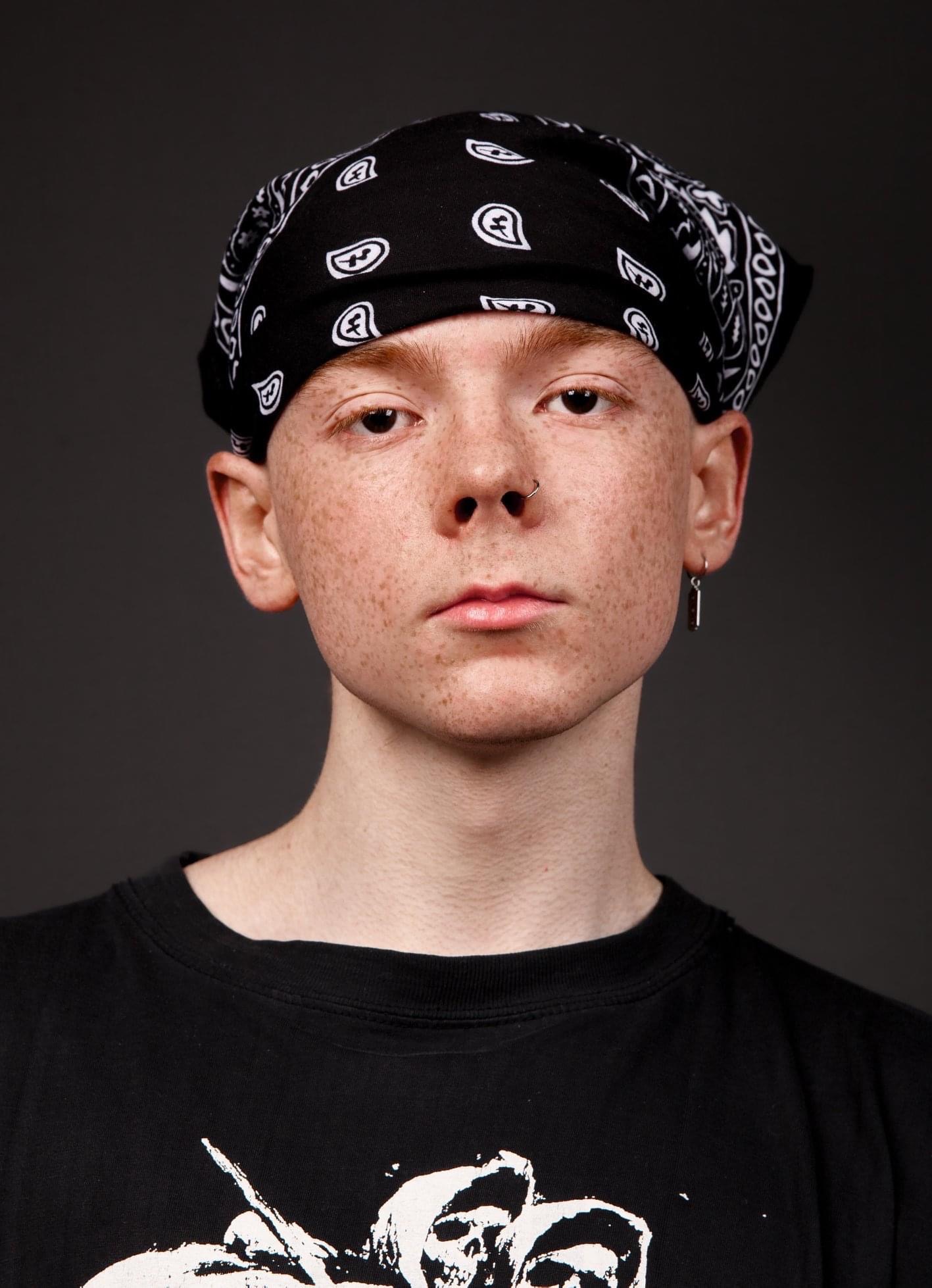 Portret młodego mężczyzny z piegami, w czarnej bandanie z białym wzorem, kolczykiem w nosie i podłużnym kolczykiem w uchu, na ciemnoszarym tle.