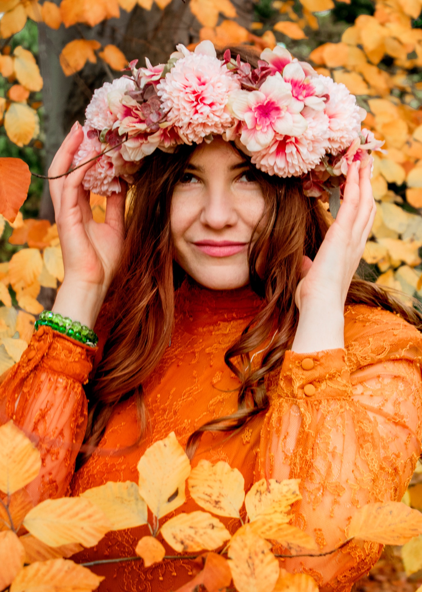 Portret kobiety z rudymi włosami, ubranej w pomarańczową koronkową sukienkę i wianek z różowych kwiatów na głowie, pozującej na tle jesiennych liści.