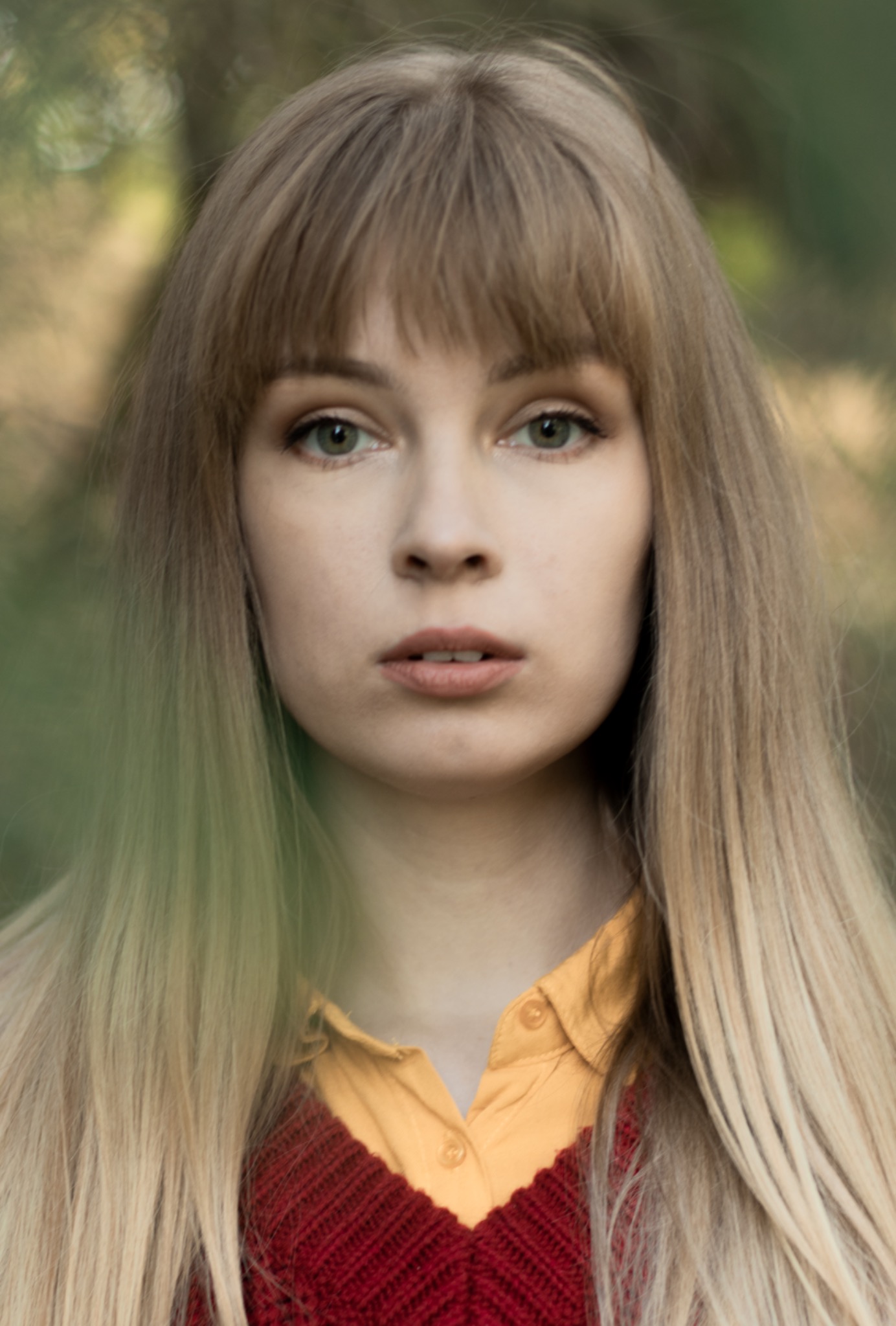 Portret młodej kobiety z prostą grzywką i długimi, blond włosami z ombre, ubrana w żółtą koszulę i bordowy sweter na tle zamazanej zieleni.