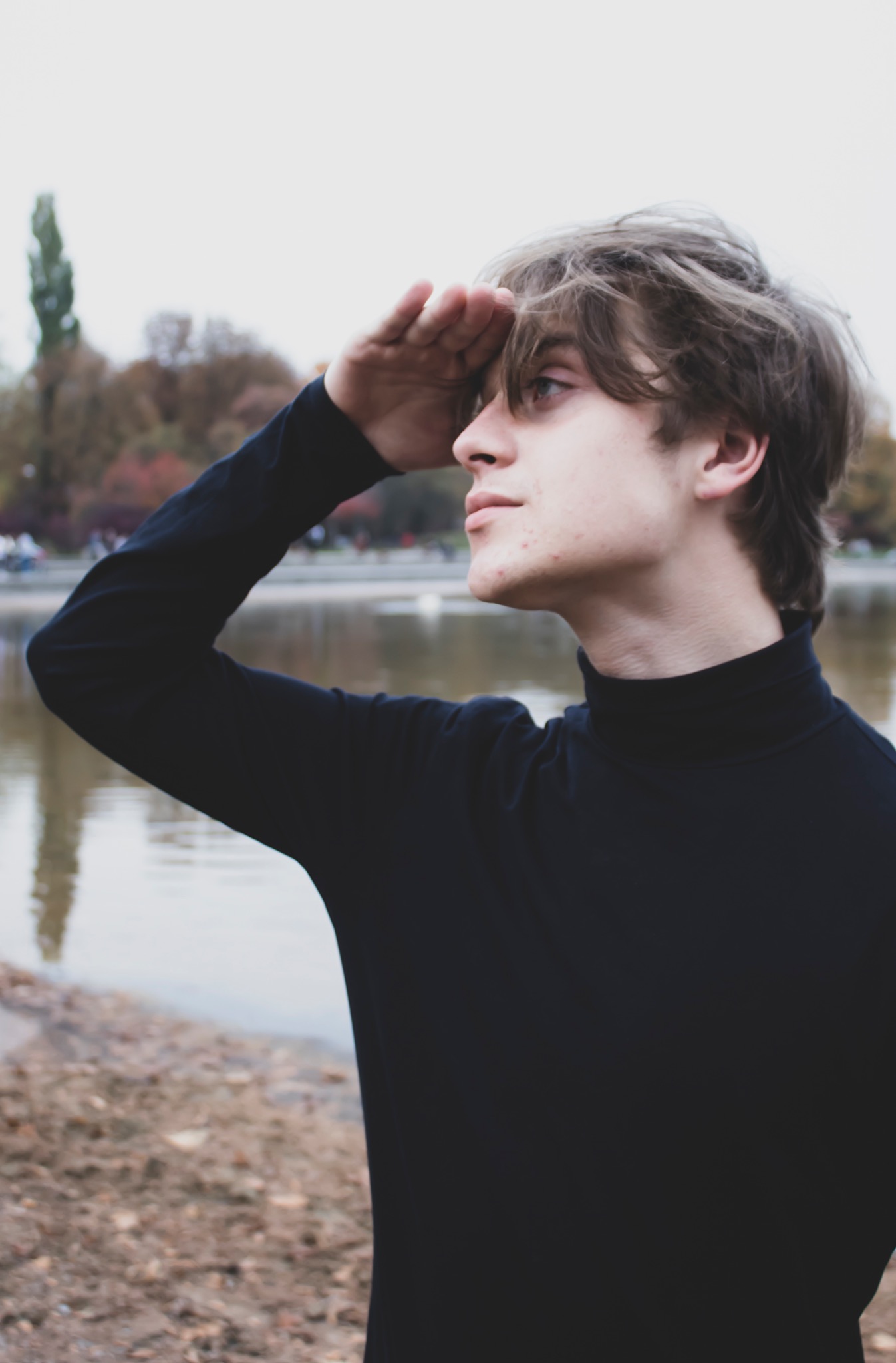 Młody mężczyzna w czarnym golfie osłania oczy dłonią, patrząc w dal na tle jeziora i parku jesienią.