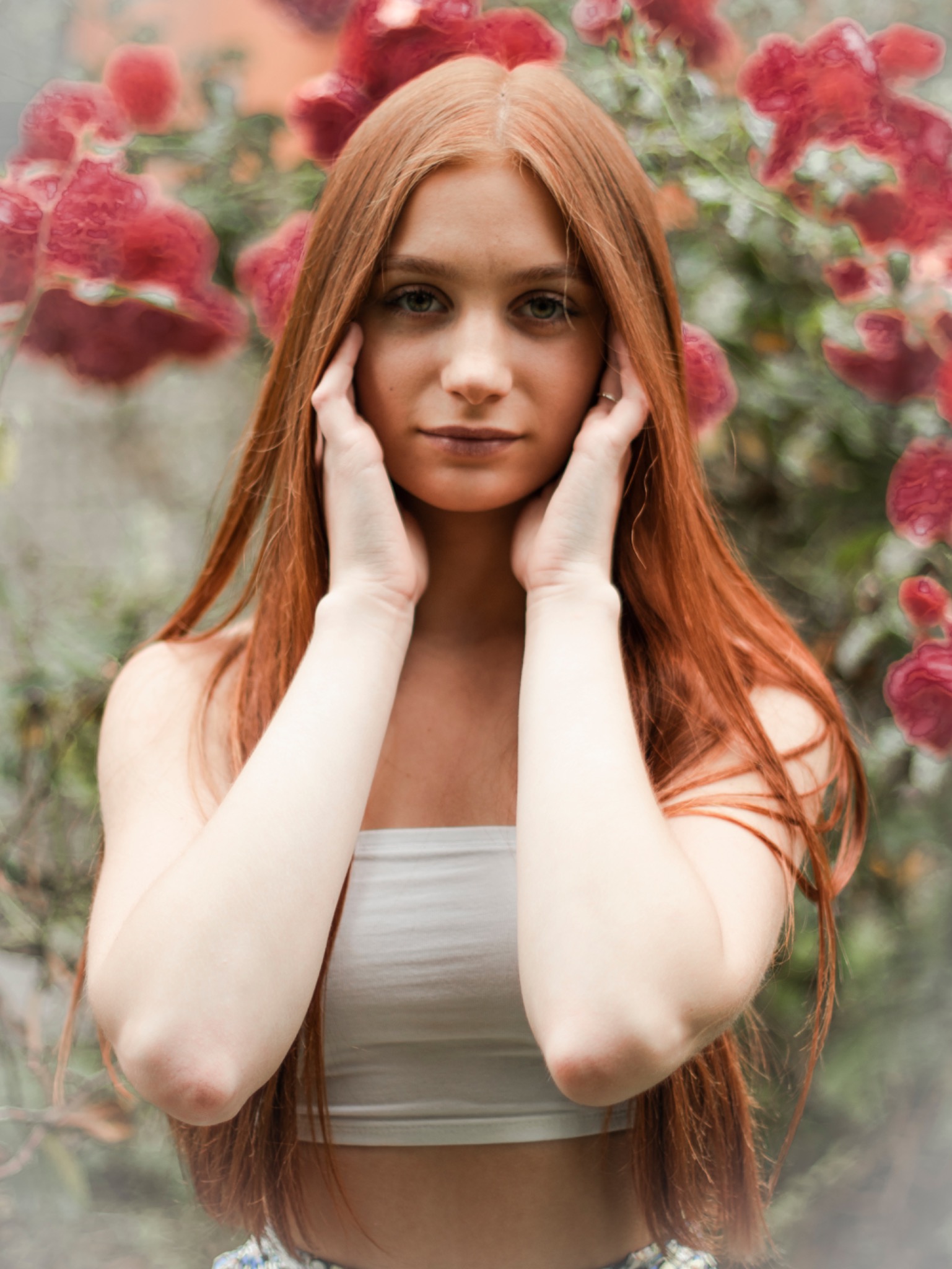 Portret młodej kobiety z długimi, rudymi włosami, delikatnie dotykającej twarzy dłońmi na tle rozmytych czerwonych róż i zieleni.