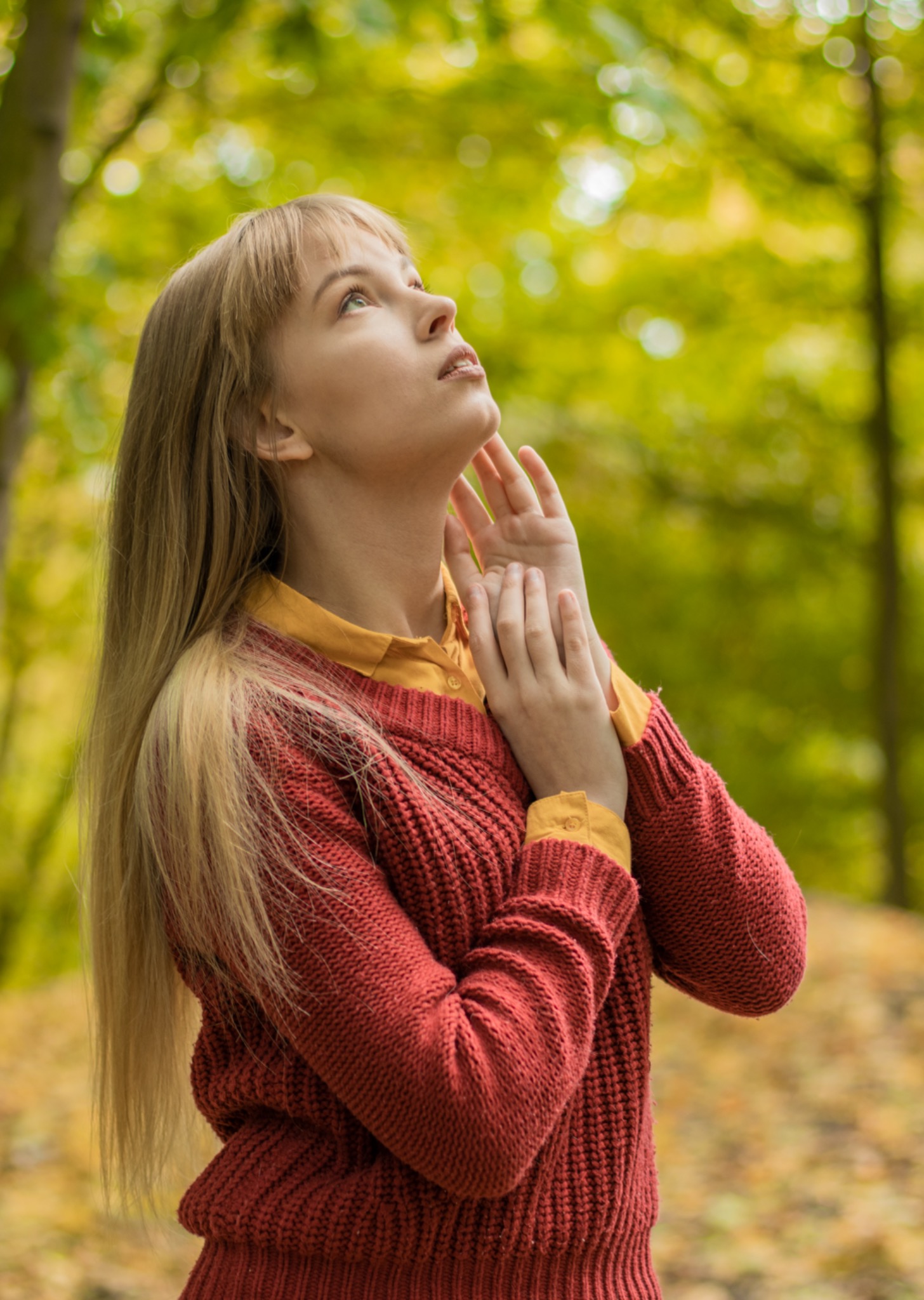 Portret młodej kobiety z blond włosami, spoglądającej w górę z rękami uniesionymi do twarzy na tle jesiennego lasu.