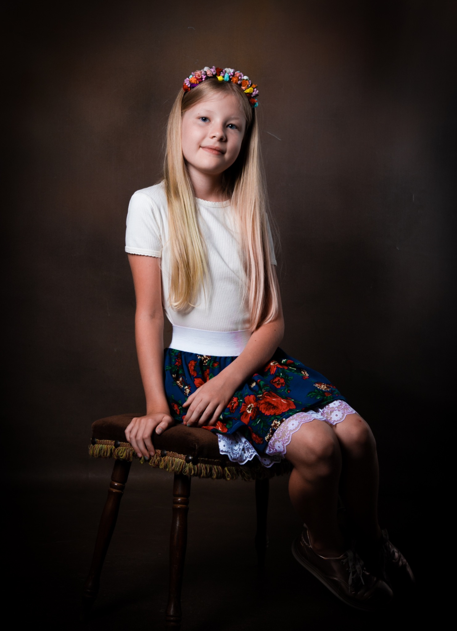 Portret młodej dziewczyny z blond włosami, siedzącej na brązowym stołku, w kwiecistej spódnicy z koronką i wiankiem na głowie, na ciemnym tle.