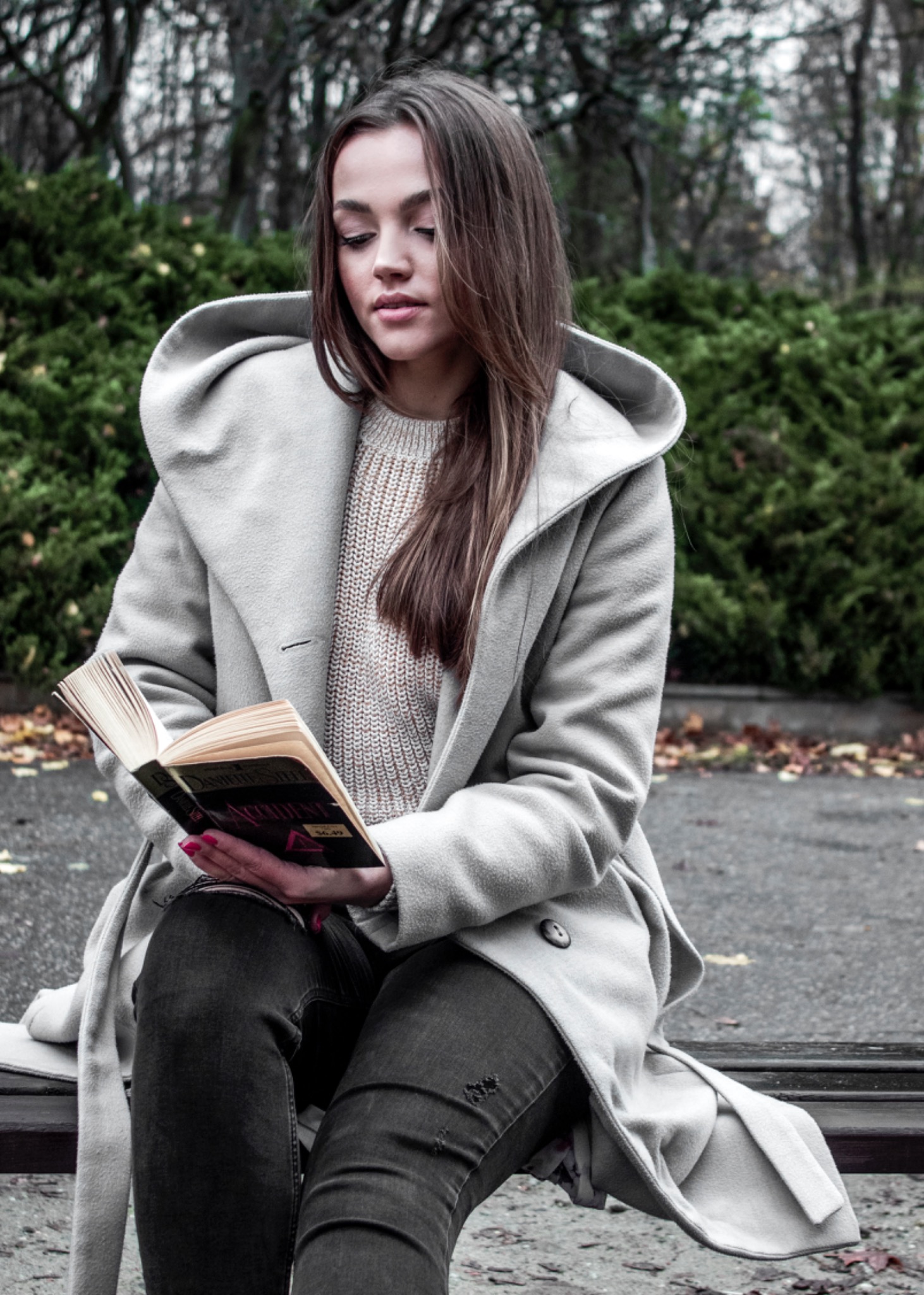 Kobieta w jasnym płaszczu z kapturem siedzi na ławce w parku, czytając książkę z okładką w odcieniach czerni i czerwieni, z manicure w kolorze fuksji.