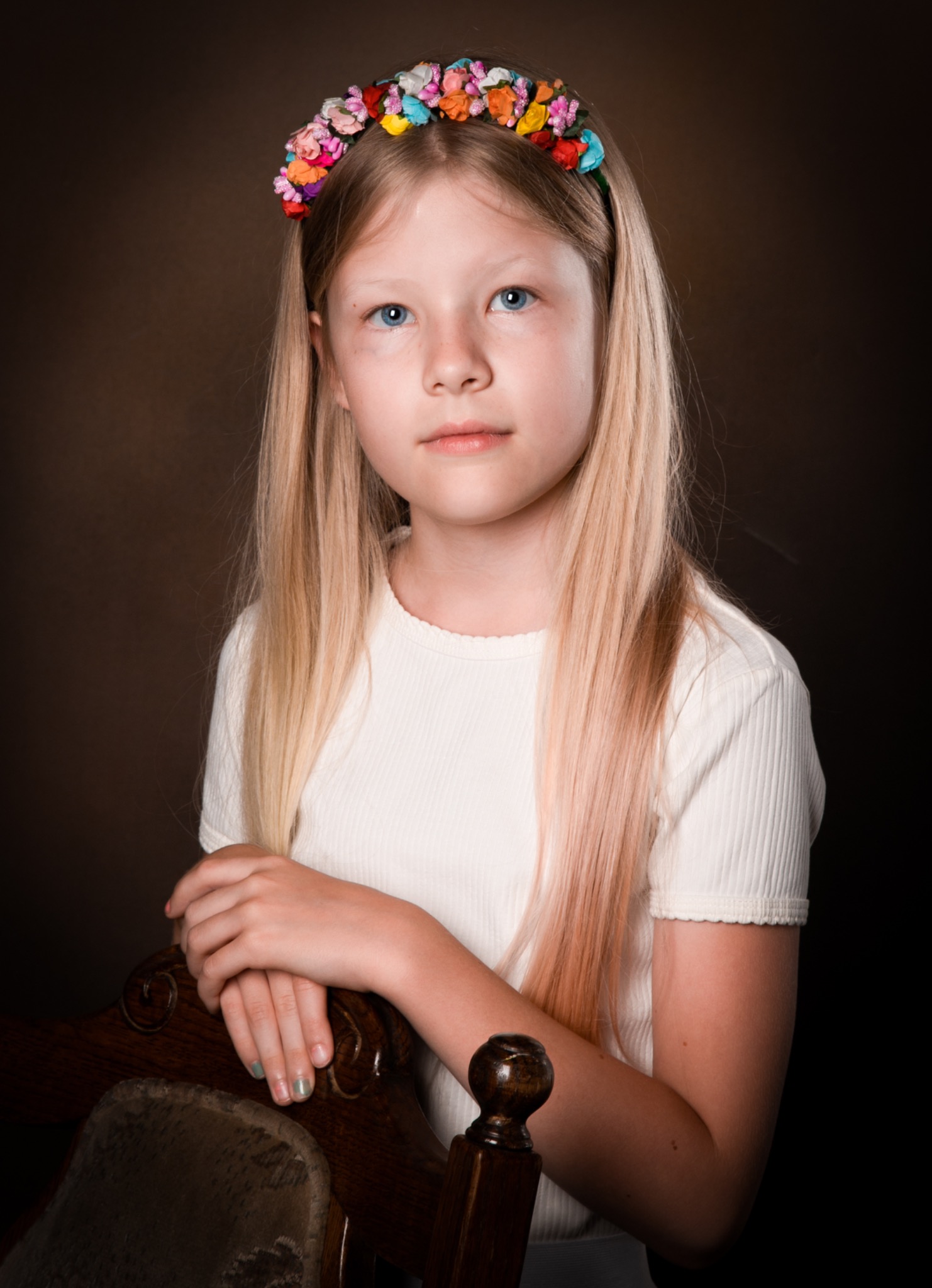 Portret dziewczynki z blond włosami i wiankiem z kolorowych kwiatów na głowie, opierającej ręce na oparciu drewnianego krzesła na ciemnym tle.