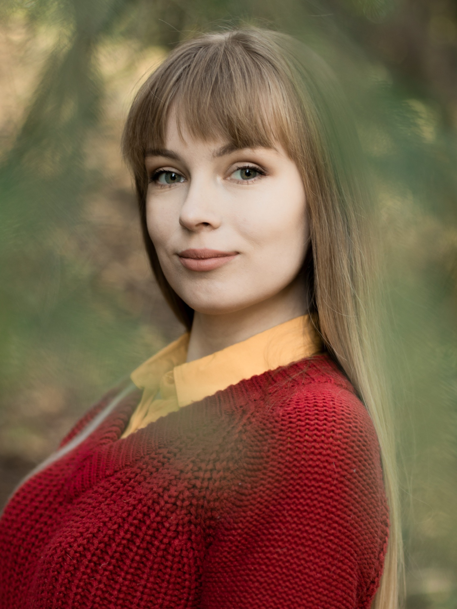 Portret młodej kobiety z długimi blond włosami, w czerwonym swetrze i żółtej koszuli, na rozmytym tle leśnym.