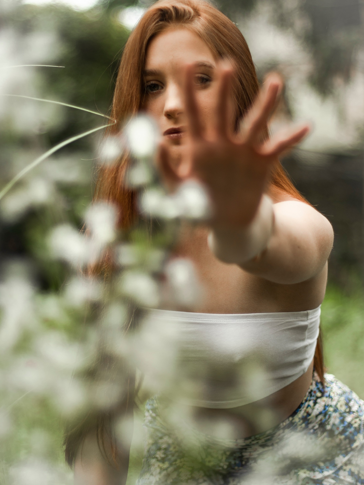 Portret kobiety z rudymi włosami, wyciągnięta dłoń w kierunku obiektywu, rozmyte kwiaty na pierwszym planie, naturalne światło.