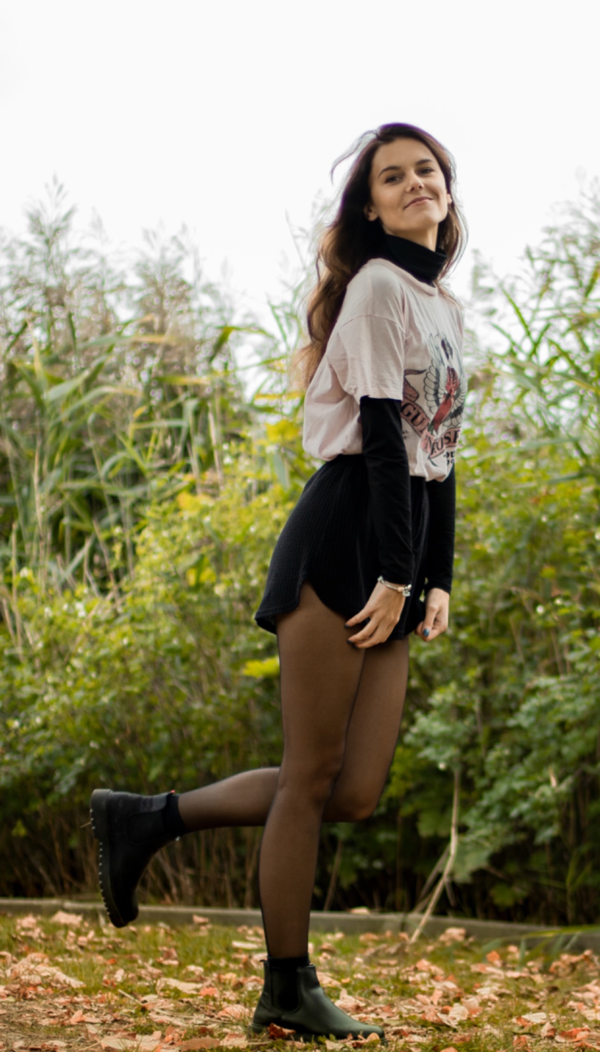 Portret młodej kobiety w jesiennej stylizacji, pozującej na tle trzcin i opadłych liści, z uniesioną nogą i delikatnym uśmiechem.