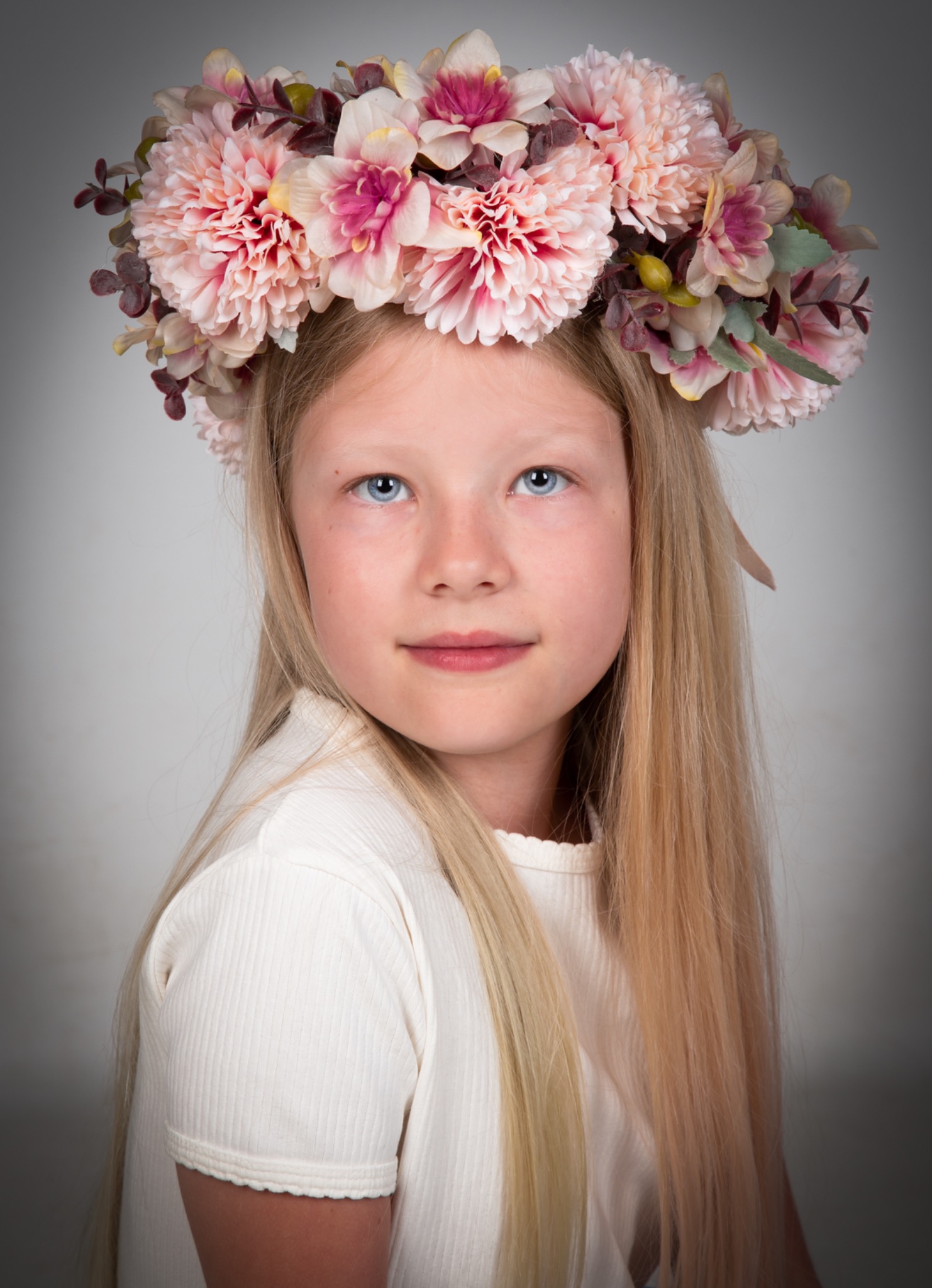 Portret młodej dziewczyny z długimi blond włosami i wiankiem z różowych i białych kwiatów na głowie, spoglądającej w górę na jasnym tle.