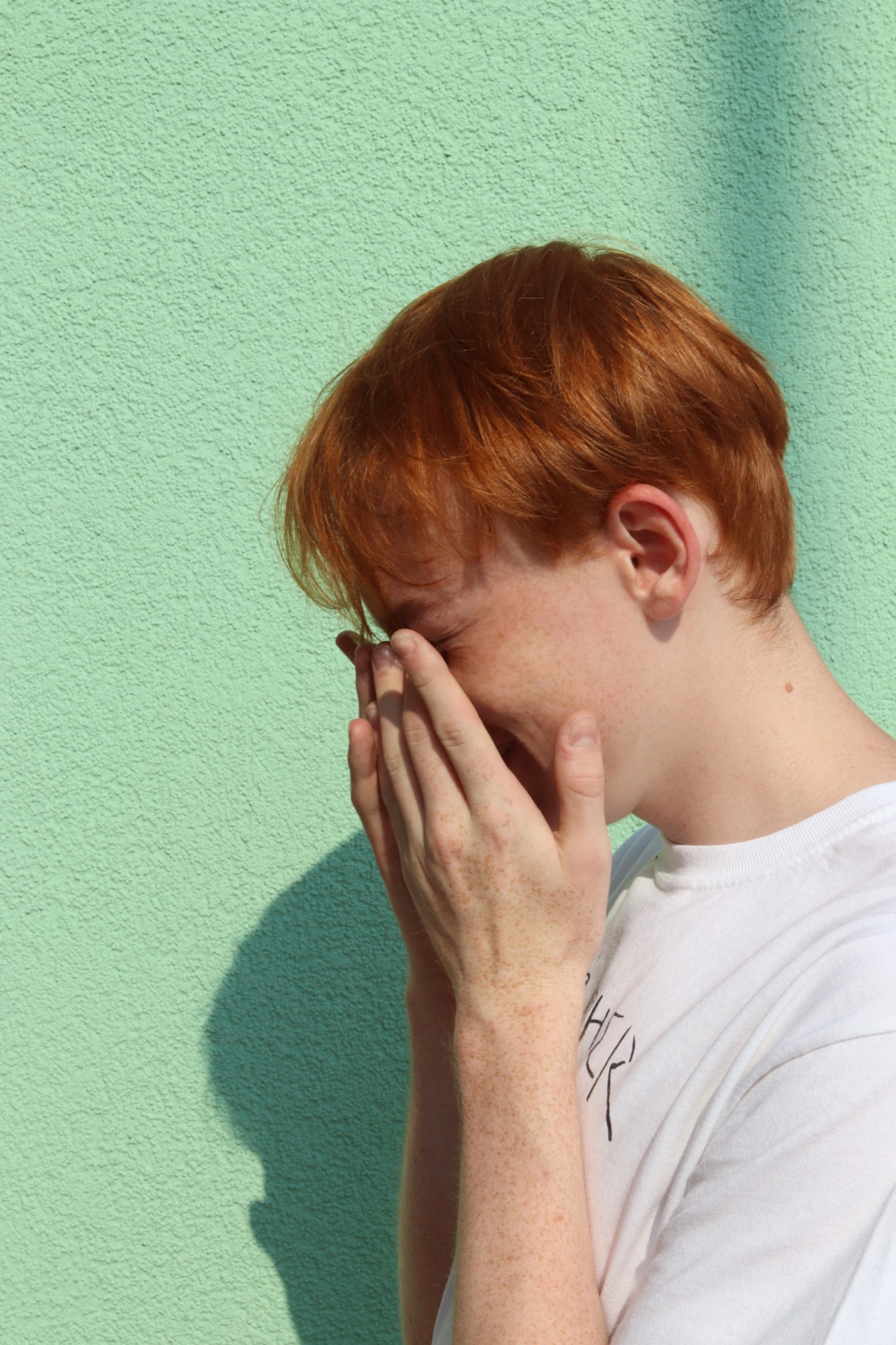 Młody mężczyzna z rudymi włosami i piegami zakrywa twarz dłońmi na tle zielonej ściany w słoneczny dzień.