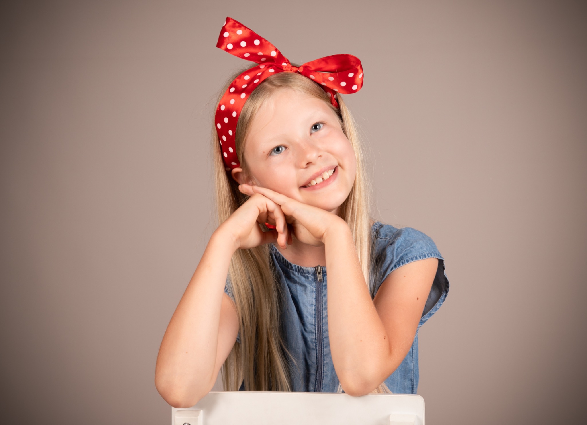 Uśmiechnięta blondynka z niebieskimi oczami, w dżinsowej sukience i czerwonej opasce w białe kropki, opiera podbródek na dłoniach, siedząc na białym krześle.