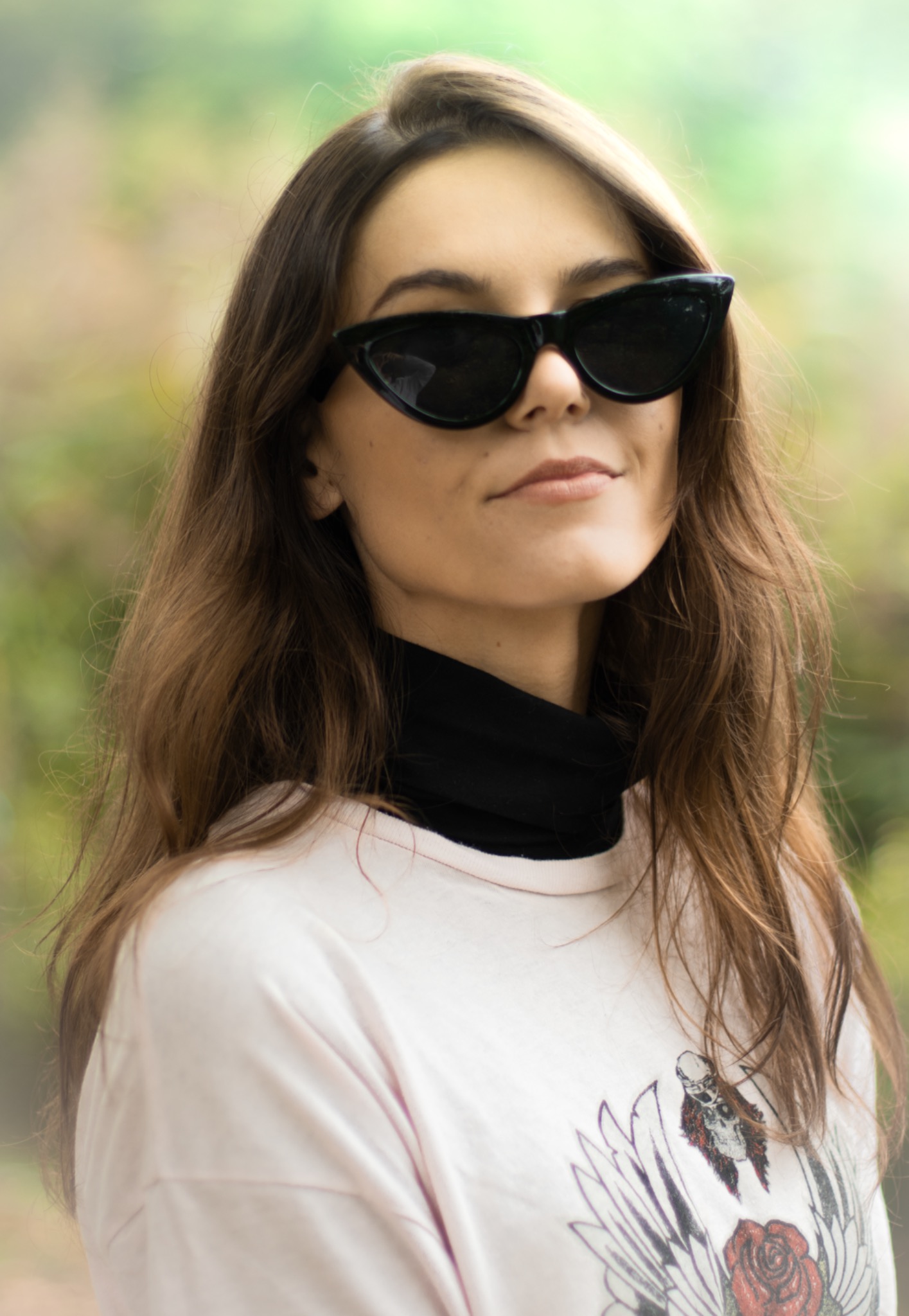 Portret kobiety w okularach przeciwsłonecznych typu kocie oko, ubrana w różową bluzę z nadrukiem, na rozmytym tle zieleni.